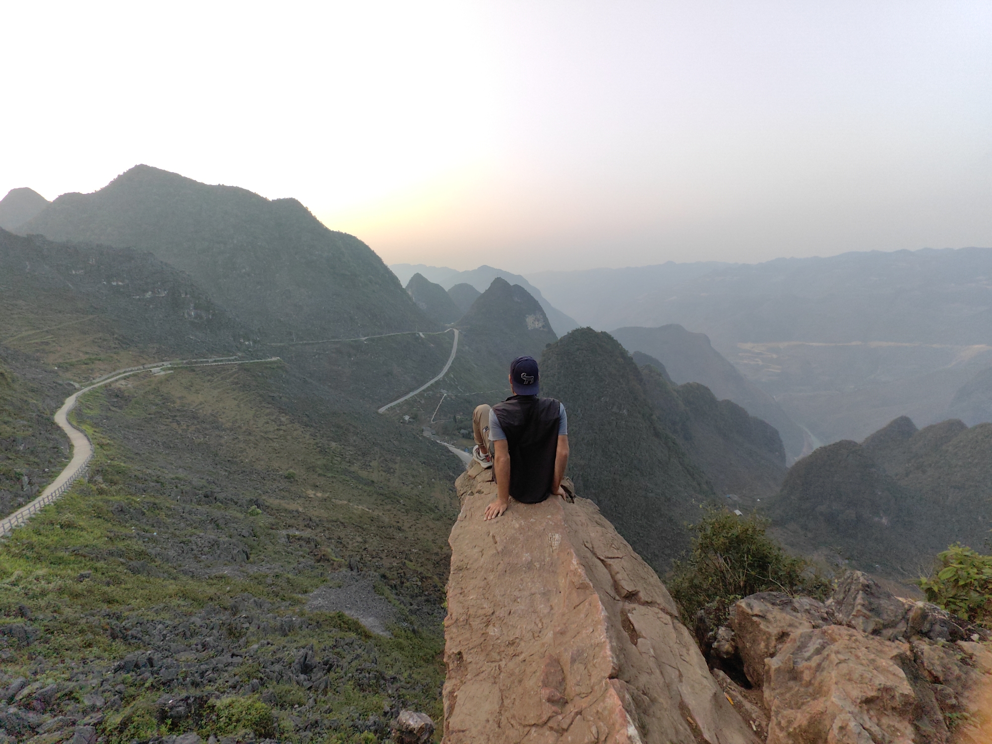 A man sits on the edge of a rock formation in the Đồng Văn Commune, Vietnam, facing away from the viewer. He is wearing a blue baseball cap, a black vest, light brown pants, and white sneakers. He looks out over a mountainous landscape. The terrain consists of steep, green-gray mountains. A winding road is visible on the left side of the image. The sky is light gray.