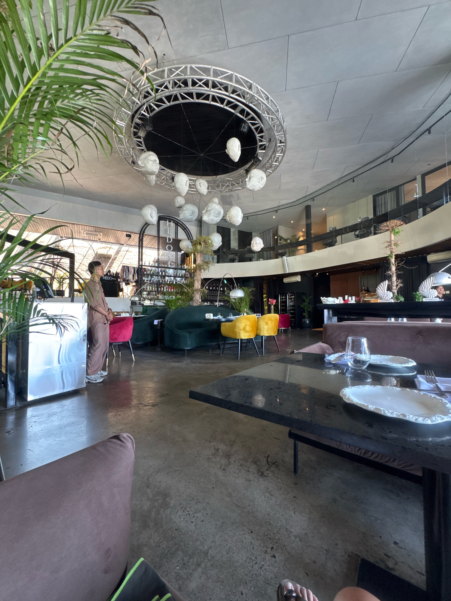 The image depicts the interior of a modern restaurant in Kuta Selatan, Indonesia. The scene is captured from a low angle, looking across a black, polished table set with white plates and glasses. In the background, a man in a light brown uniform stands near a silver counter, appearing to be a staff member. The restaurant features a circular mezzanine level, numerous bottles lining shelves behind the bar area, and a distinctive circular light fixture with hanging white, head-like sculptures. Lush green plants, including a large palm in the foreground, add to the ambiance. Seating includes plush green and red chairs, along with yellow velvet chairs. The overall atmosphere is upscale and contemporary.