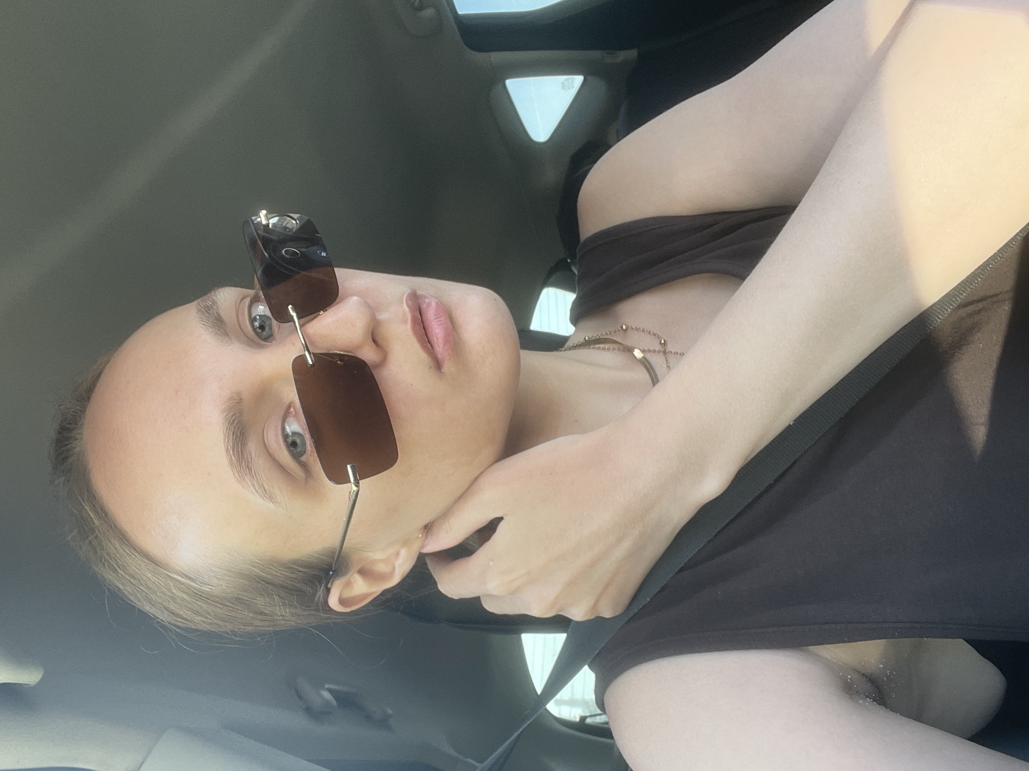 A woman wearing sunglasses and a brown tank top is sitting in a car. She has fair skin and light-colored hair. Her hand is near her face. The car has a sunroof. The location is not evident in the image to determine whether it is in Izhevsk, Russia.