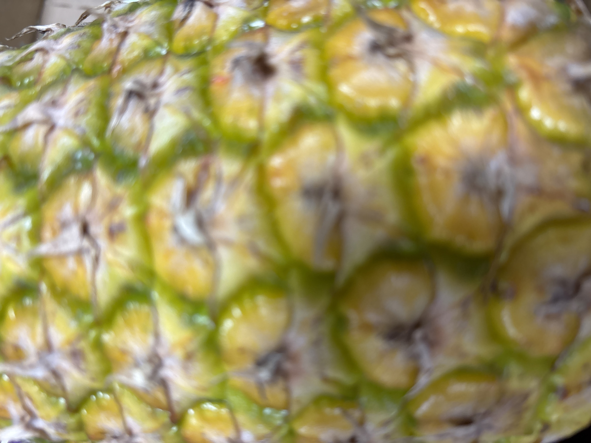 This is a close-up, out-of-focus photograph of the textured skin of a pineapple. The image predominantly features the characteristic diamond-shaped segments of the pineapple's rind, with variations of yellow and green hues, some appearing lighter and more golden, others a deeper, almost olive green. The texture is rough and scaly, with visible lines and small brown or grey markings on some of the segments. The lighting creates highlights and shadows, emphasizing the uneven surface. There is no discernible background or context that would place this image in Zvenigorod, Russia, or any specific city. The focus is entirely on the surface of the fruit.