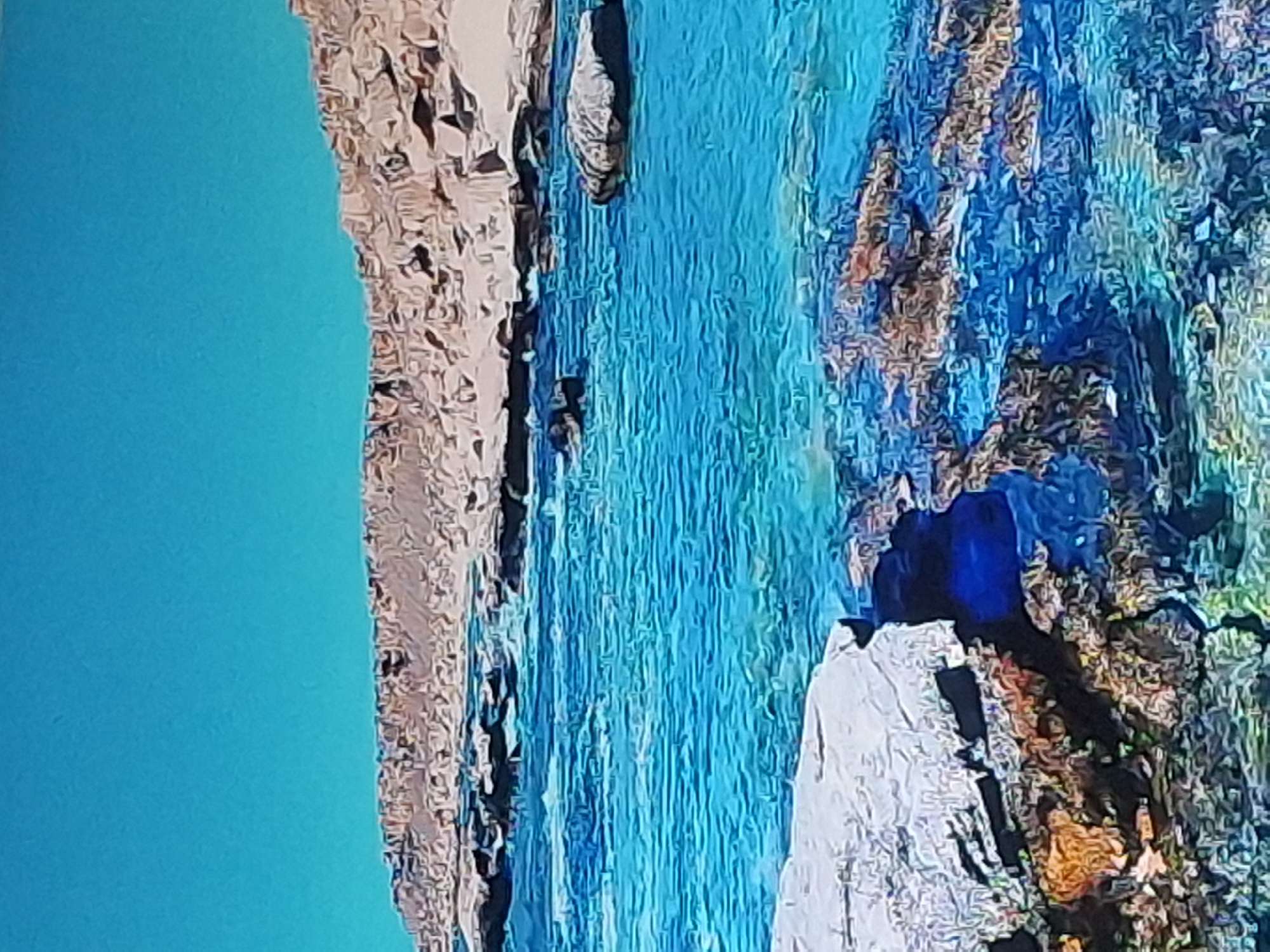 The image depicts a coastal landscape with clear blue water and a rocky shoreline under a bright blue sky. The foreground features a large, light-colored rock with an area of dark water or shadow beneath it, adjacent to submerged rocks with brown and orange hues. The water is turquoise and shows gentle ripples. In the middle ground, the water is deeper blue with visible waves and scattered rocks. The background shows a rocky, elevated landmass with a light brown and tan color, possibly a cliff or hillside. The overall scene conveys a natural, serene environment. The image is not associated with the city of Izmail, Ukraine.