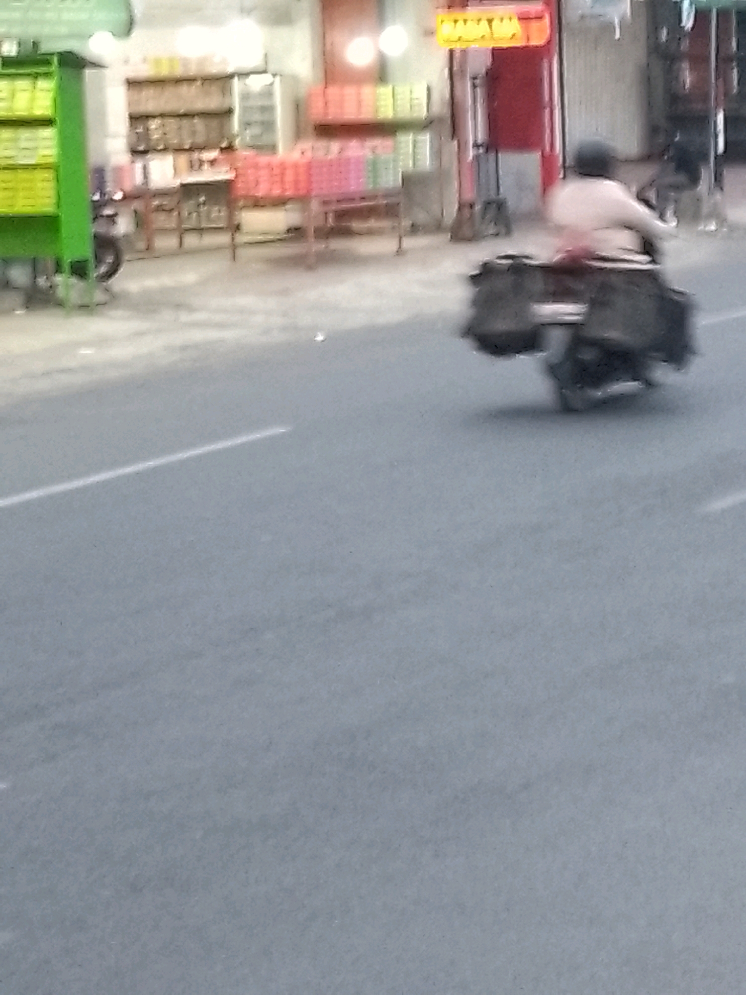 In Jember, Indonesia, a person on a motorcycle is captured in motion, with the image's blur indicating significant movement. The rider is wearing a helmet and is seated on the motorcycle, which is equipped with saddlebags on either side, suggesting a journey or transport of goods. In the background, a storefront is visible, with shelves stocked with various items, possibly packaged goods or supplies. The storefront has a bright, illuminated sign with yellow lettering, and a red facade. The motorcycle and rider are on a paved road, with a white line marking a lane. The overall scene depicts a typical street view in a commercial area of Jember.