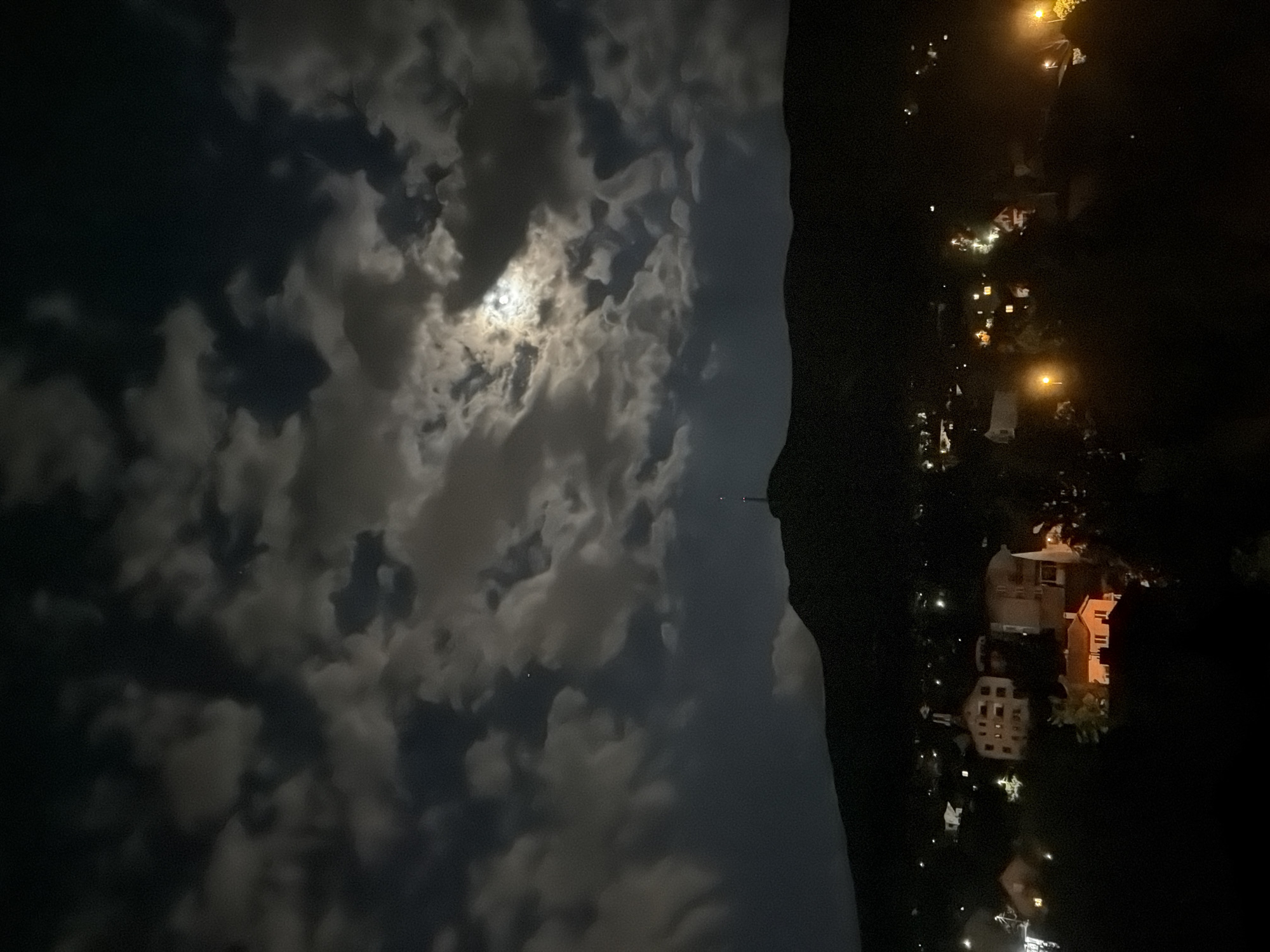 The image is a nighttime shot of a cloudy sky over a residential area. The moon is visible through the clouds. On the right side of the image, a dark hillside separates the sky from a cluster of houses and streetlights below. This location appears to be in Sofia, Bulgaria, due to the style of architecture and landscape. There are no people visible in the image. The focus is on the contrast between the illuminated clouds and the darkness of the night.