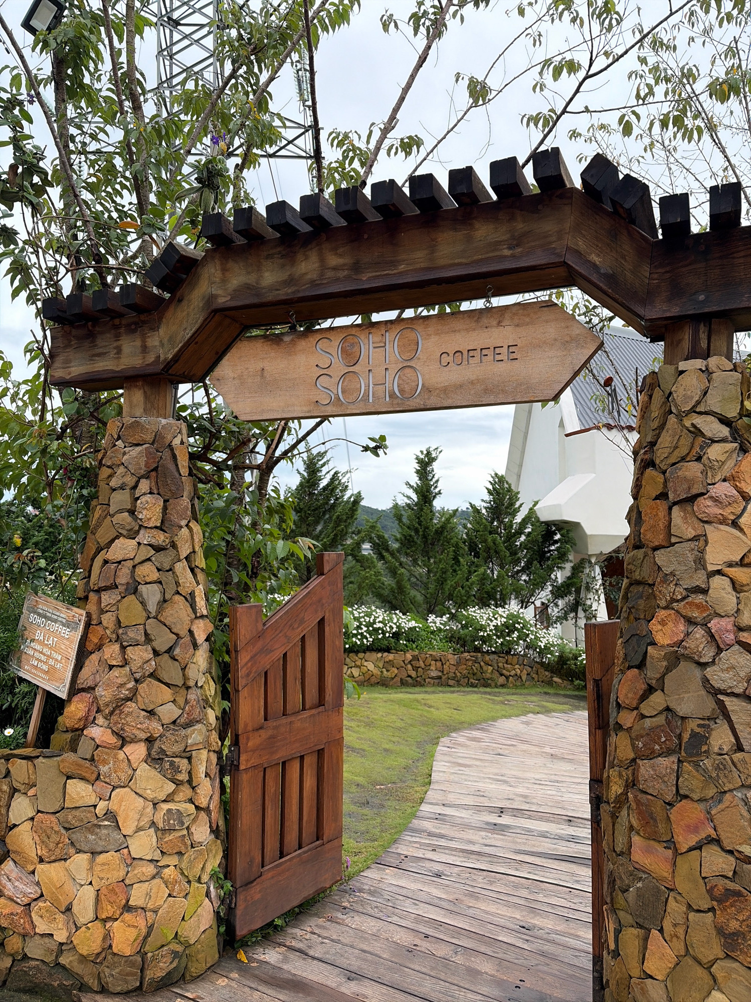 The image shows the entrance to "SOHO COFFEE" in Da Lat, Vietnam. The entrance features a rustic wooden archway supported by stone pillars. A wooden sign on the archway reads "SOHO COFFEE" with an arrow pointing to the right. A wooden gate is open, revealing a pathway made of wooden planks that curves into a grassy area. In the background, a white building with a distinctive roofline is visible, surrounded by greenery and trees. A small sign on the left pillar displays the address and operating hours of the coffee shop: "SOHO COFFEE DA LAT 21 HOÀNG HOA THÁM P. XUÂN HƯƠNG - ĐÀ LẠT. LAM DONG." The scene suggests a tranquil, scenic location in Da Lat, a city known for its cool climate and natural beauty.