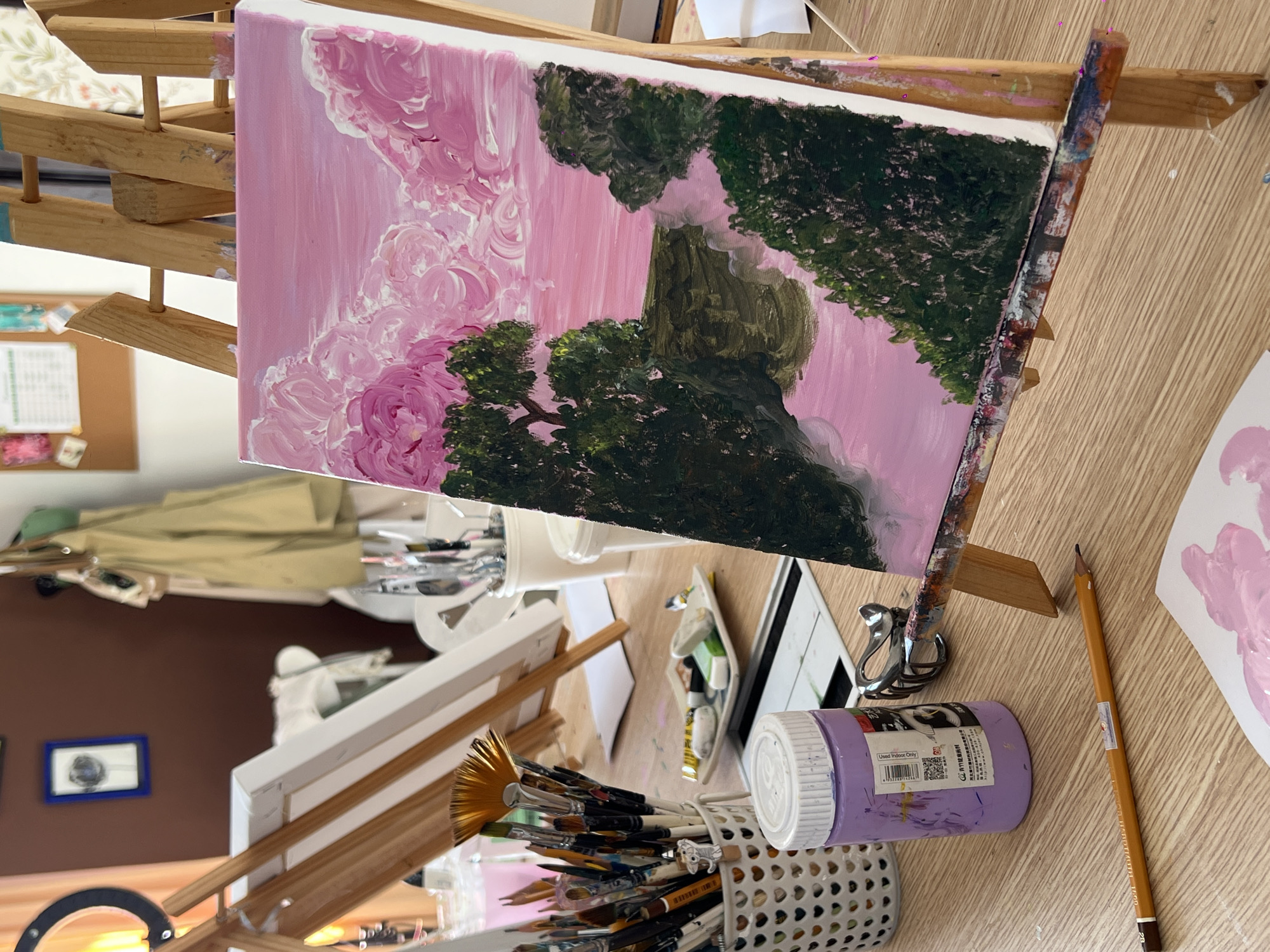 This is an indoor, eye-level, close-up shot of an art studio setup. The main subject is a partially finished painting of a landscape with pink clouds and green trees, displayed on a wooden easel. The painting is rendered in a loose, impressionistic style. In the foreground, a purple jar of paint and a pencil lie on a wooden table. To the left, a white basket holds a collection of paintbrushes. In the background, another easel with a blank canvas is visible, along with art supplies and tools. The overall impression is of a creative space dedicated to painting. The image does not contain any explicit geographical markers that would suggest a specific location within Bac Nha Trang Ward, Vietnam.