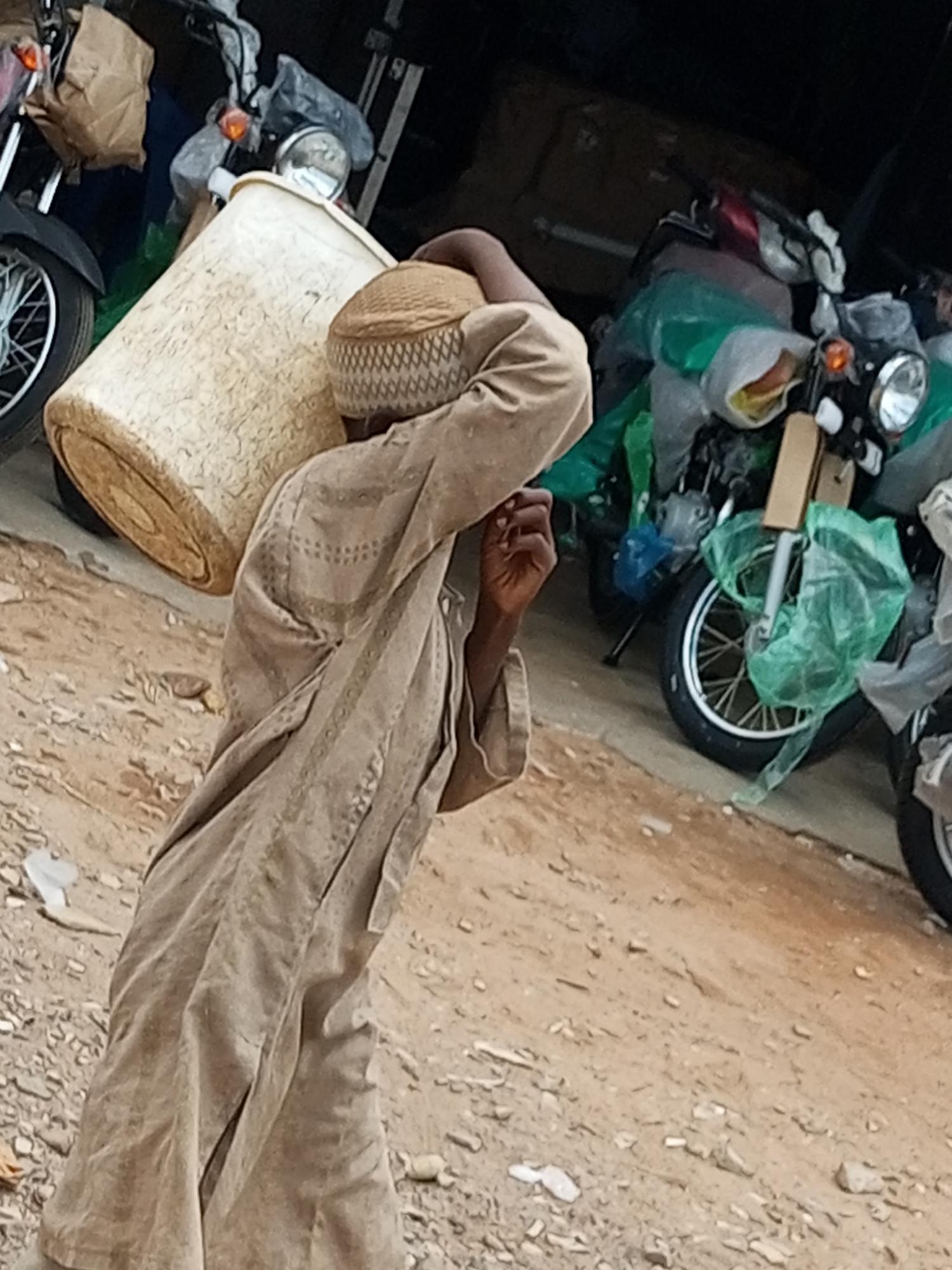 A person is carrying a large, pale, cylindrical object on their shoulders. The person is wearing a light brown, loose-fitting garment and a patterned cap. The object appears to be a woven basket or container with a textured surface. The scene takes place outdoors on a dirt surface with scattered small stones and debris. In the background, several motorcycles are visible, some partially covered in green plastic. One motorcycle in the foreground has its front wheel and part of its frame showing. The lighting suggests it is daytime. This scene is consistent with a market or storage area in the city of Gombe, Nigeria, where it is common to see people carrying goods and motorcycles being used for transport and sale.