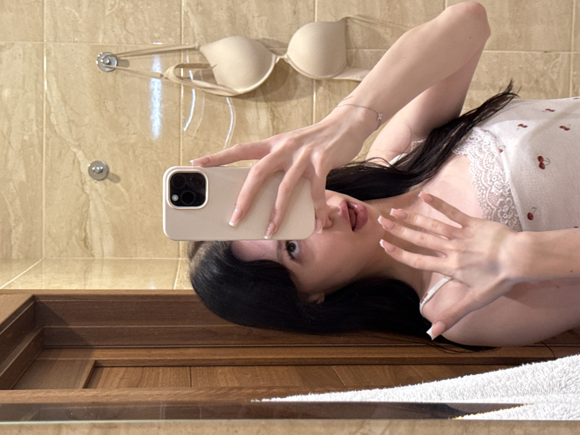 A young woman is taking a mirror selfie in a bathroom. She has long dark hair and is wearing a white camisole with a cherry pattern and lace trim. She is holding a light-colored smartphone in her right hand, capturing her reflection. Her left hand is raised, with fingers splayed. Her left arm has a thin silver bracelet with a butterfly charm. In the background, beige marble tiles line the walls, and a wooden door is visible to the left. A beige bra hangs from a hook on the wall behind her. A white towel hangs on the left side of the frame. The image appears to be taken indoors in a residential bathroom.