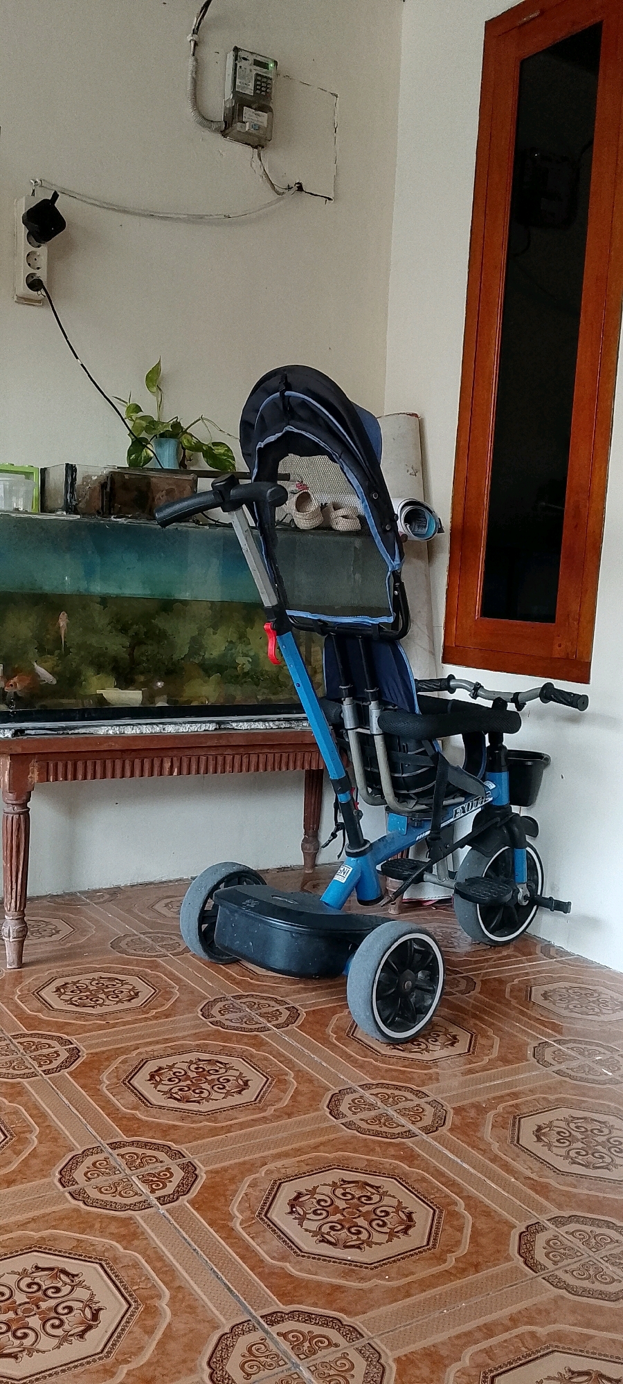 This is a medium shot, eye-level, indoor photograph taken in Satriamekar, Indonesia. The scene is a tiled floor with ornate brown and tan patterned tiles. A blue and black stroller with three wheels is positioned in the center of the frame. The stroller has a canopy and a handlebar with grips. To the left of the stroller is a wooden table with a glass-topped aquarium filled with green plants and fish. Behind the aquarium, on a white wall, is an electrical meter box and a power outlet with a plug. To the right of the stroller is a wooden-framed window with dark glass. The lighting is natural and casts soft shadows. There are no people present in the image.