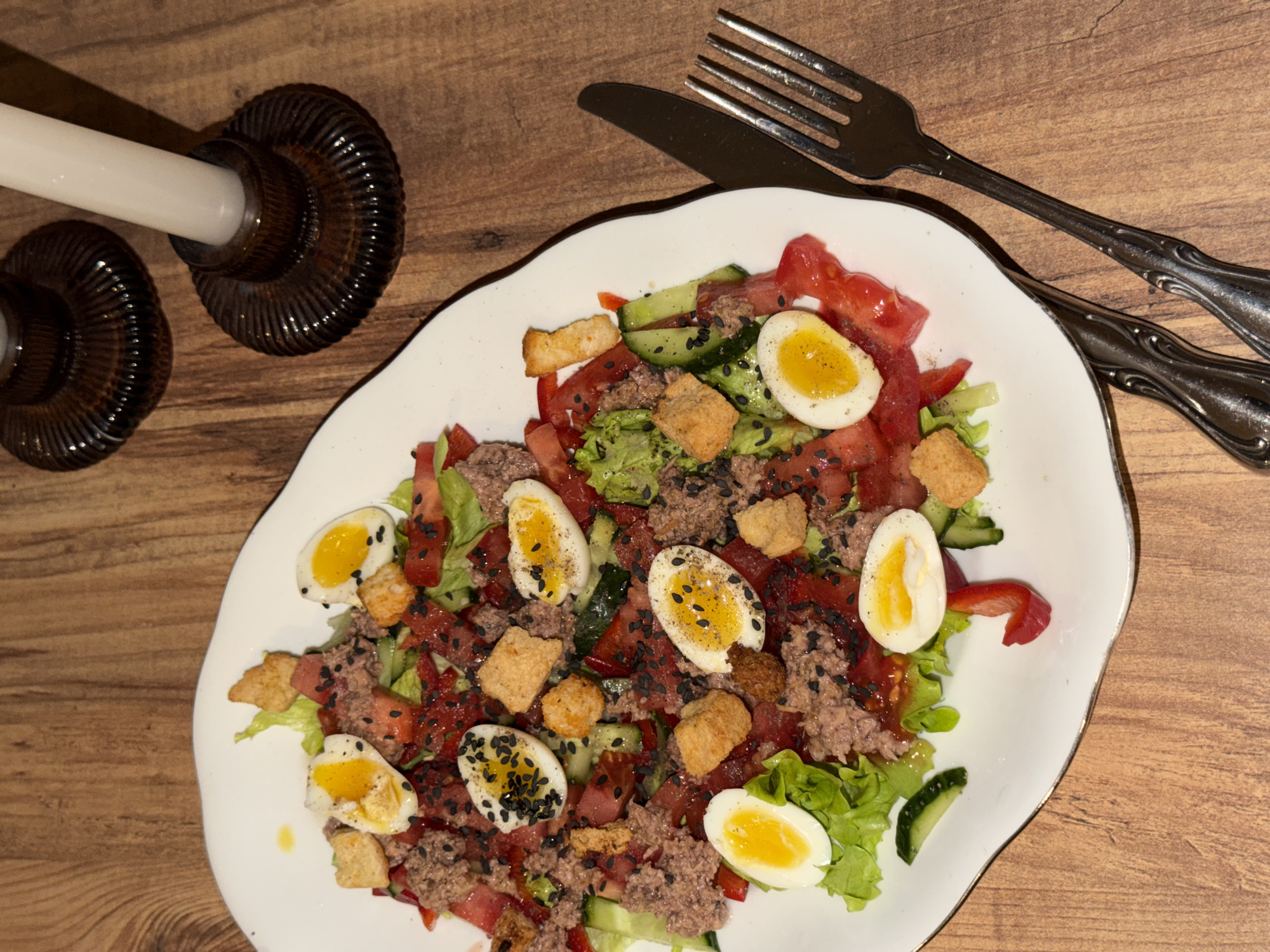 On a wooden table, a white, irregularly shaped plate is arranged with a salad. The salad contains lettuce, diced tomatoes, cucumber slices, croutons, and shredded meat, possibly tuna. Halved hard-boiled eggs, some sprinkled with black seeds, are distributed throughout the salad. To the left of the plate, there are two short, dark-colored candle holders made of a ribbed material, each holding a white candle. To the right of the plate, a fork and knife are positioned. The scene is not explicitly identifiable as being in Tbilisi, Georgia.