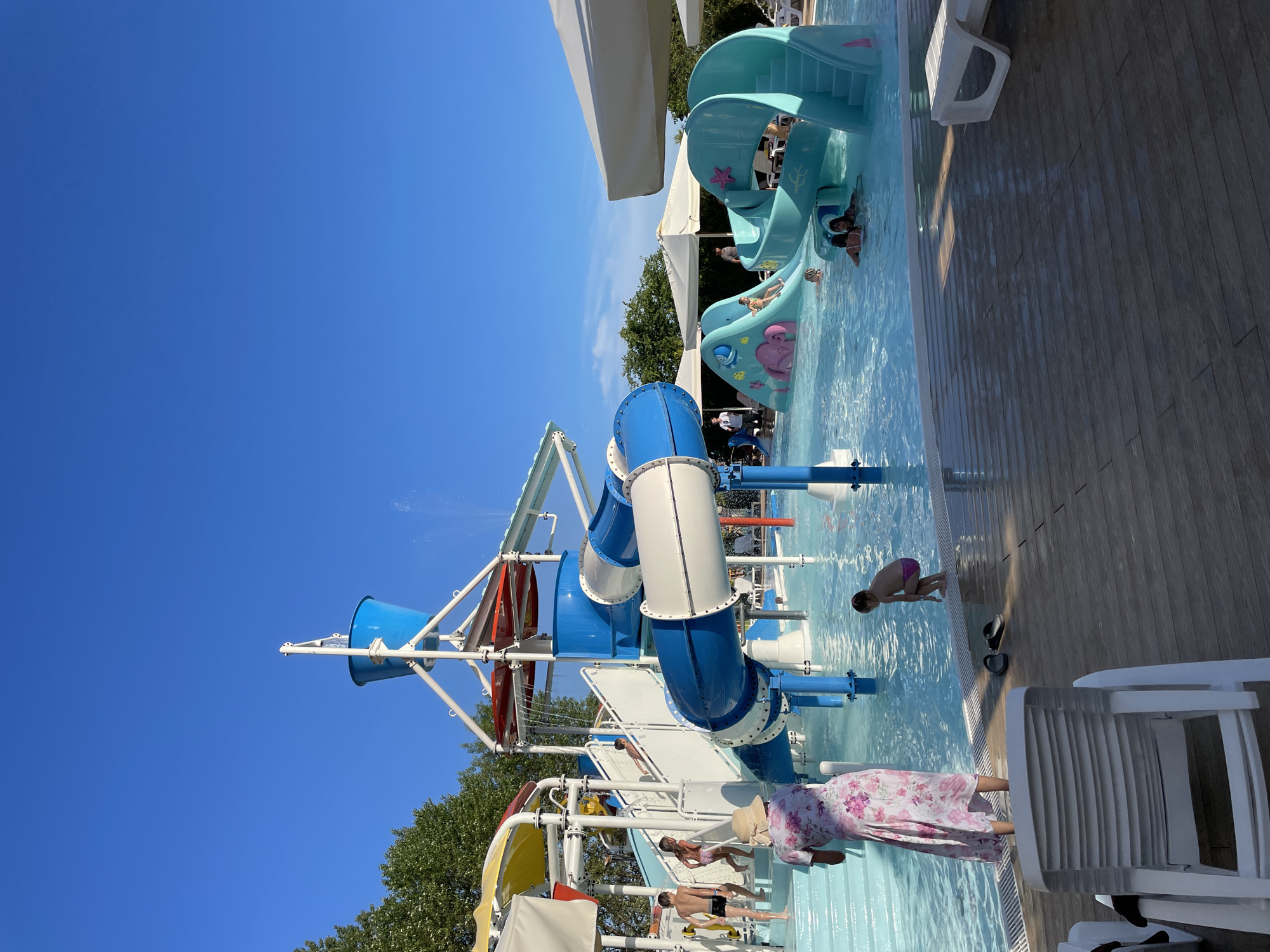 This image depicts a sunny day at an outdoor water park in Sochi, Russia. The main focus is a large, multi-level water slide complex with bright blue and white tubing, and a large blue tipping bucket that is currently empty. Children are visible playing in the shallow pool surrounding the slides. To the right, there is a smaller, teal-colored water slide structure with playful sea creature designs. Several white umbrellas are set up around the pool area, providing shade. In the foreground, a woman in a floral dress stands by the pool, and a white plastic lounge chair is visible on the wooden deck. The clear blue sky indicates fair weather.
