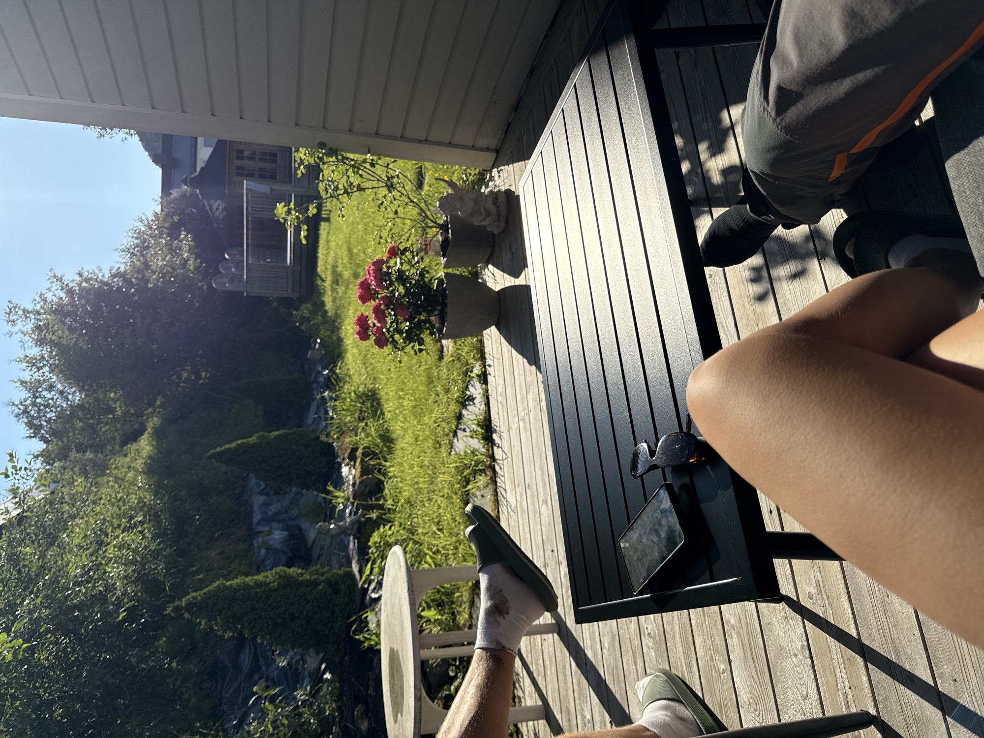 The image depicts a sunny outdoor scene, likely a backyard or patio, with a wooden deck in the foreground. A black slatted table occupies the center of the frame, with a smartphone and sunglasses resting on its surface. Two individuals are partially visible, one on the left with a leg in a green slide sandal and white sock resting on a round table, and another on the right with a leg in dark pants with an orange stripe.
Behind the deck, a well-maintained lawn is illuminated by sunlight. A cluster of bright pink flowers in a pot is visible on the deck, alongside a small stone statue of an angel. Further back, a wooden gazebo with a dark roof stands on a raised platform, with trees and foliage forming a natural backdrop. A light blue sky with subtle sunlight flares is visible above. The presence of manicured landscaping and a gazebo suggests a residential setting. Given the context, this scene could be located in the city of Harstad, Norway, which is known for its scenic natural beauty and well-kept outdoor spaces.