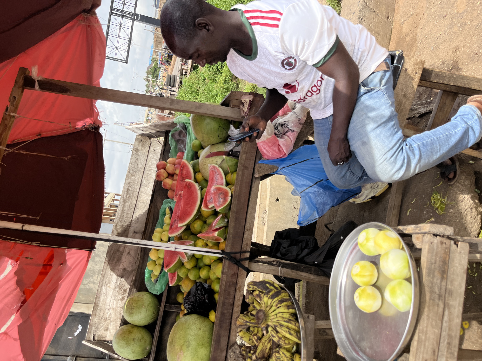 In Bukuru, Nigeria, a man sits at a fruit stall under a red awning. He is wearing a white t-shirt with green trim and light blue jeans. He is looking at his phone. In front of him is a wooden cart piled high with various fruits, including watermelons, oranges, and bananas. To his left, on a metal plate, are several peeled yellow fruits, possibly lemons or limes. The stall is set up on a dirt road with a background of buildings and vegetation.
