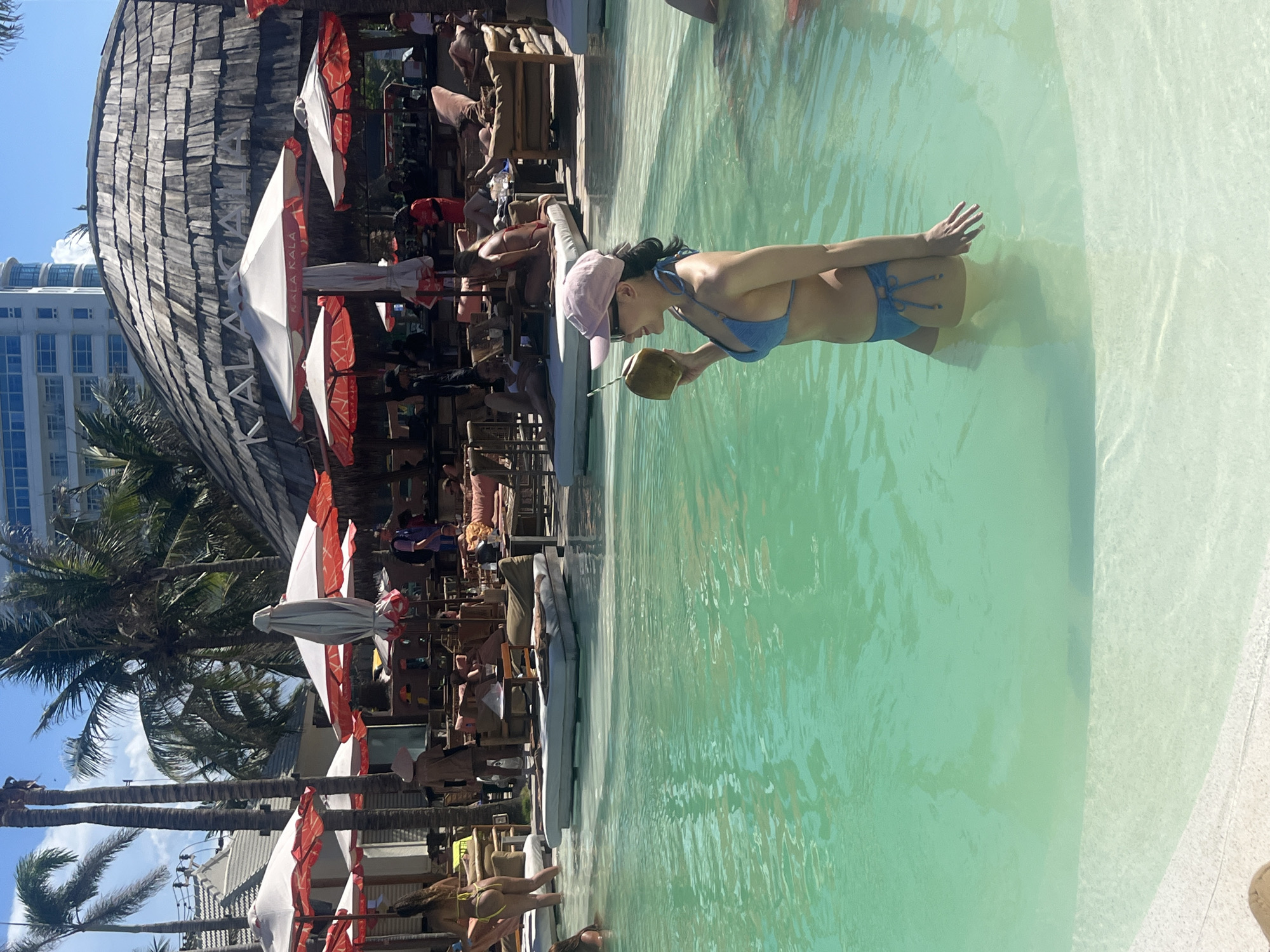 The image captures a vibrant daytime scene at a resort or beach club, set in Da Nang, Vietnam.
In the foreground, a woman wearing a light blue bikini and a pink baseball cap stands in the clear, light green-blue water of a large swimming pool. She is holding a fresh green coconut, from which she appears to be drinking, with a small straw visible. Her reflection is faintly visible in the calm water.
The background reveals a bustling poolside area. A distinctive building with a dark, textured roof, possibly made of wooden shingles, prominently features the word "KALA KALA" in large letters. Numerous white and red-orange beach umbrellas are scattered around the pool, many of which also display the "KALA KALA" branding. Beneath these umbrellas and in other shaded spots, many people are seen relaxing on lounge chairs, conversing, and enjoying the sunny weather. Some individuals are also partially visible in the pool. Tall palm trees are interspersed throughout the area, adding to the tropical ambiance. In the distant background, modern high-rise buildings stand under a clear blue sky.
The bright natural light suggests it is mid-day or early afternoon, with strong sunlight creating clear shadows. The overall atmosphere conveys a sense of relaxation and leisure at a popular tropical holiday destination.