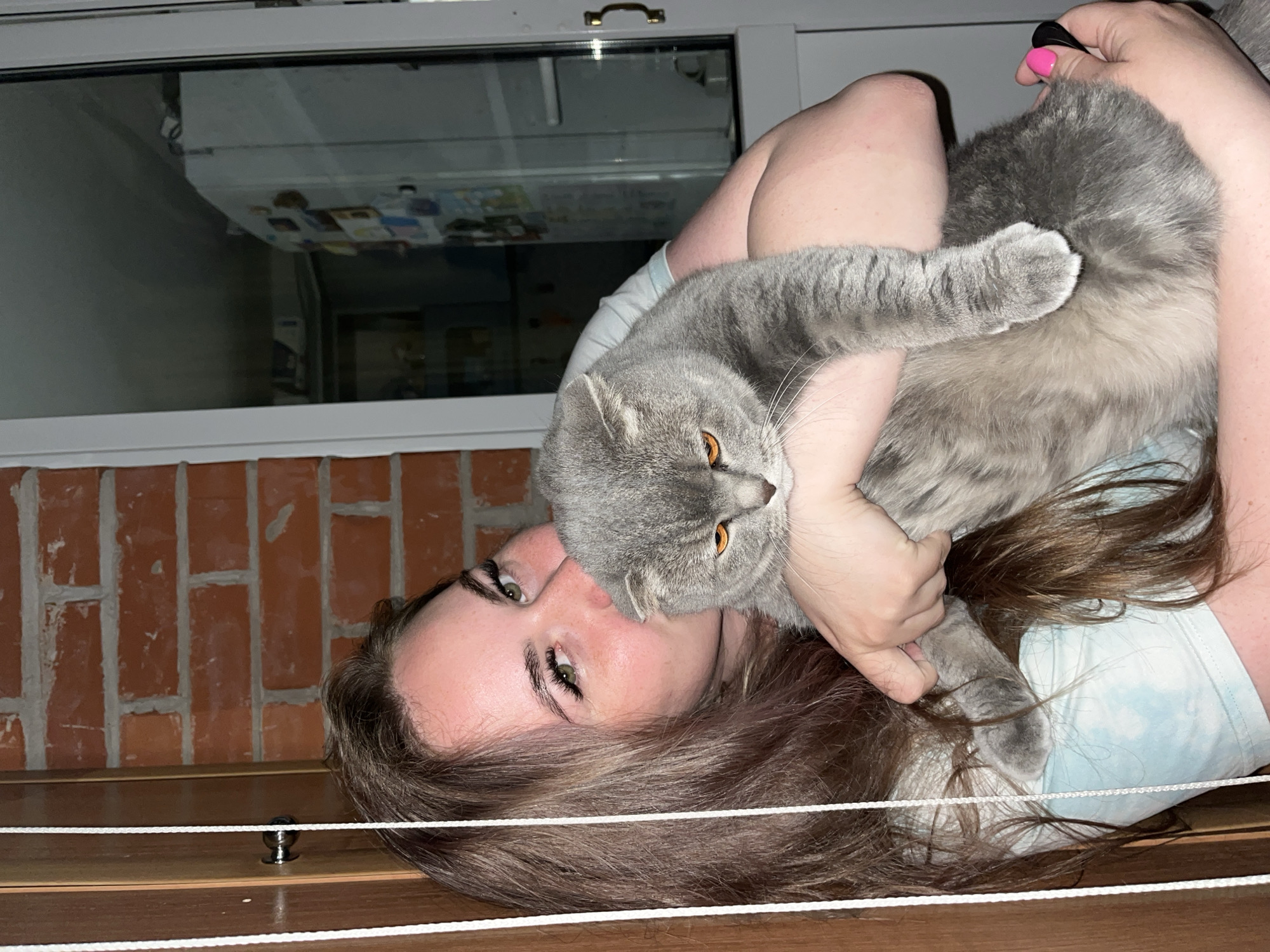 A woman lies on her back holding a gray cat. She is indoors. The cat's face is near her face, and she holds the cat with both arms. She has long, brown hair. Above her is a window with a view to what appears to be another building's brick facade. The city of Chaikovsky, Russia is not verifiable from this image.