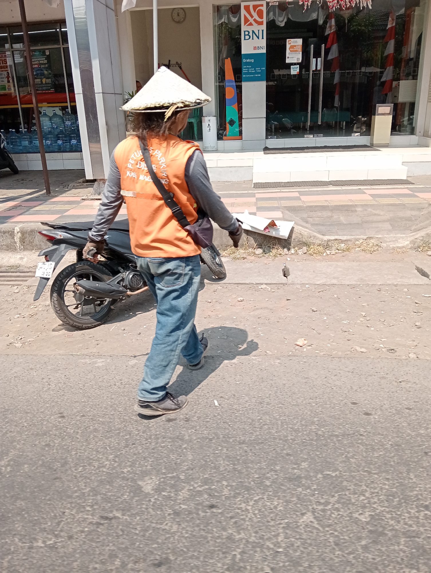 A person wearing a conical hat, an orange vest with white text, and blue jeans walks along an asphalt road. They are carrying a black bag slung over their shoulder and are walking away from the camera. A motorcycle is parked to their left. Behind the person is a building with a glass storefront displaying water bottles and advertisements. The building has the BNI logo visible. The area appears to be a commercial street in Grabag, Indonesia.