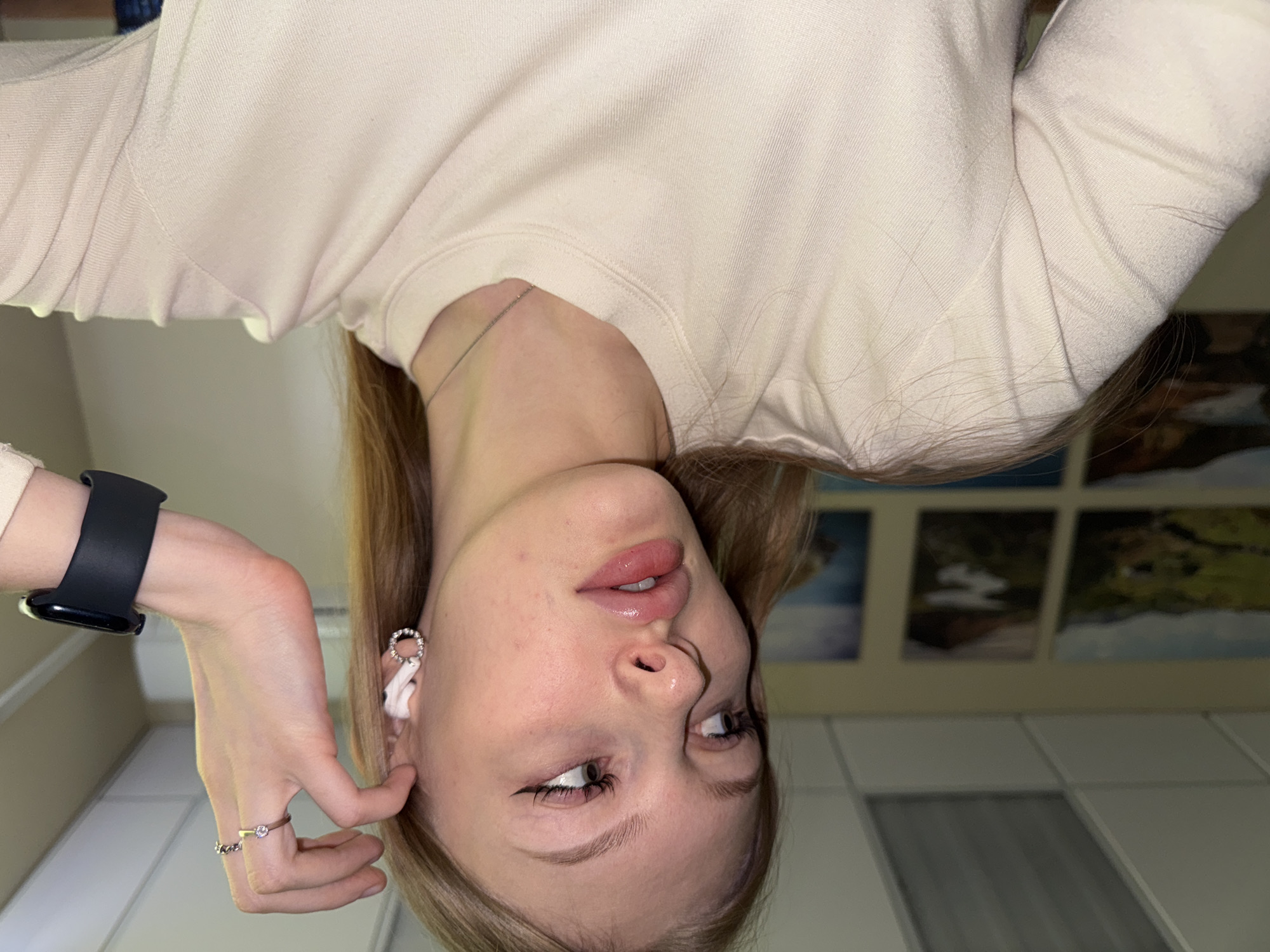The image depicts a young woman with blonde hair, fair skin, and light eyes. She is wearing a light-colored sweater and a silver chain necklace. She has an earring in her left ear, a black smartwatch on her left wrist, and rings on her left hand. She appears to be touching her left ear with her fingers. The background consists of what seems to be a wall with multiple framed pictures. Based on geolocation clues, the location is possibly in Perm, Russia, considering the style of architecture and interior design elements that align with regions in Russia. The tiles and grid-like pattern on the walls/ceiling are reminiscent of common interior finishing in Perm, Russia.