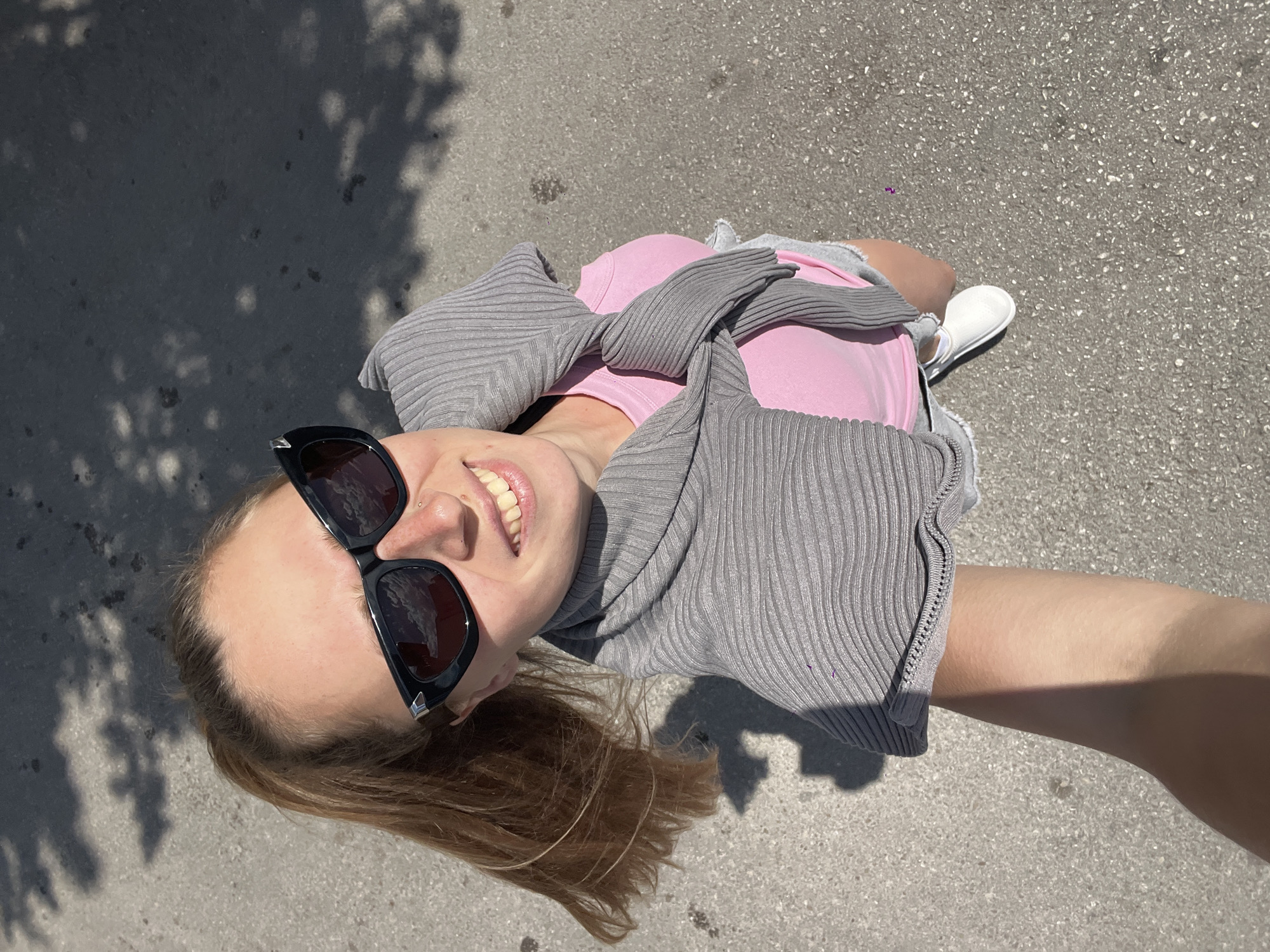 A young woman is taking a selfie outdoors in Bečići, Montenegro. She is wearing sunglasses, a pink shirt, grey sweater, and white shoes. She is standing on a paved surface that appears to be a street or walkway. Shadows from trees or foliage are visible on the ground near her head. The background shows a slightly out-of-focus view of what appears to be an urban environment.