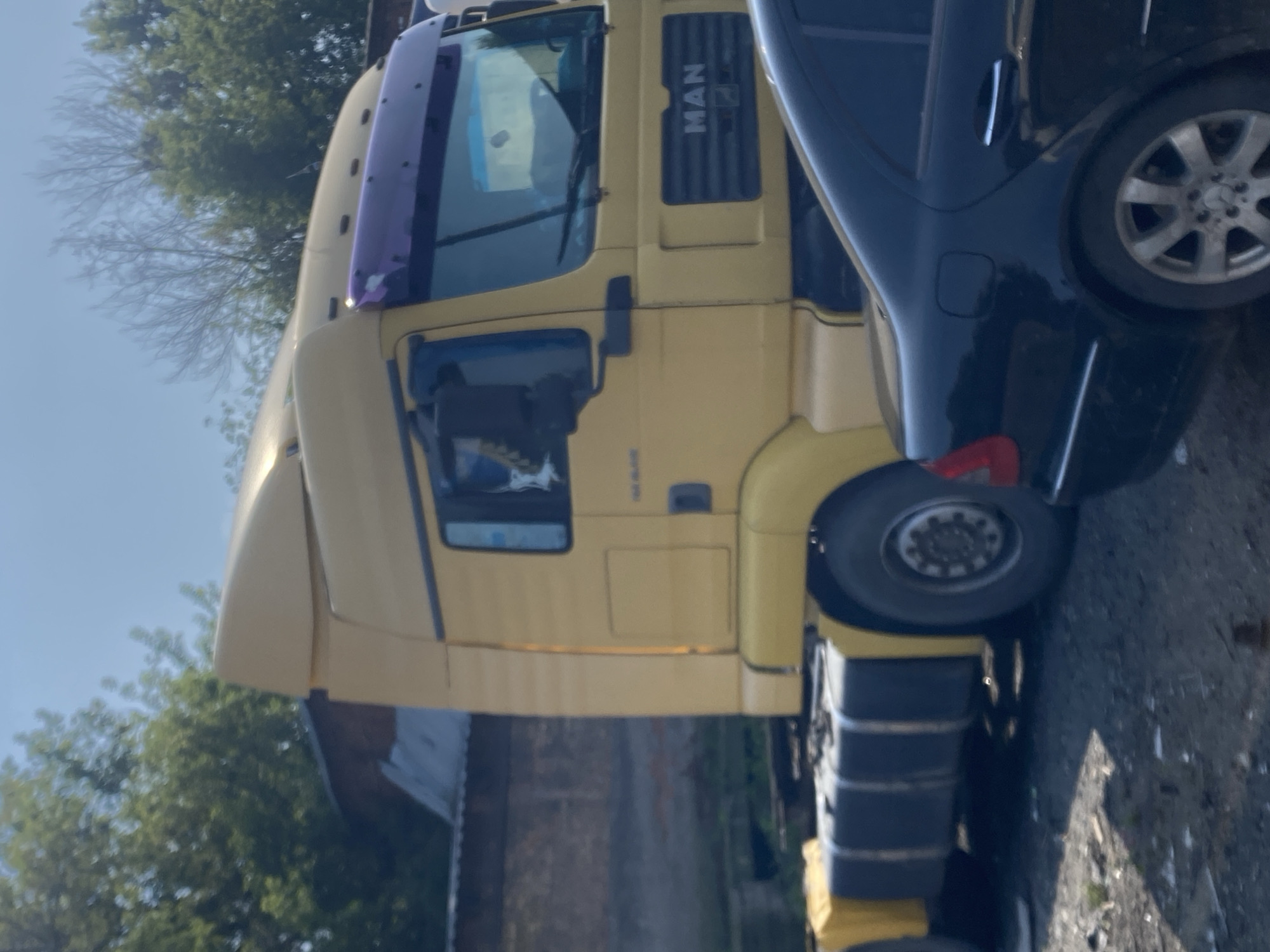 A yellow MAN semi-truck is parked outdoors, possibly in an industrial area. The truck has a large cab with tinted windows and a purple sun visor. The front grille displays the "MAN" logo. A black sedan is parked in front of the truck, partially obscuring its lower half. The background shows a brick building and trees under a clear sky. The scene is likely located in Novokuznetsk, Russia, given the implied context of the request.