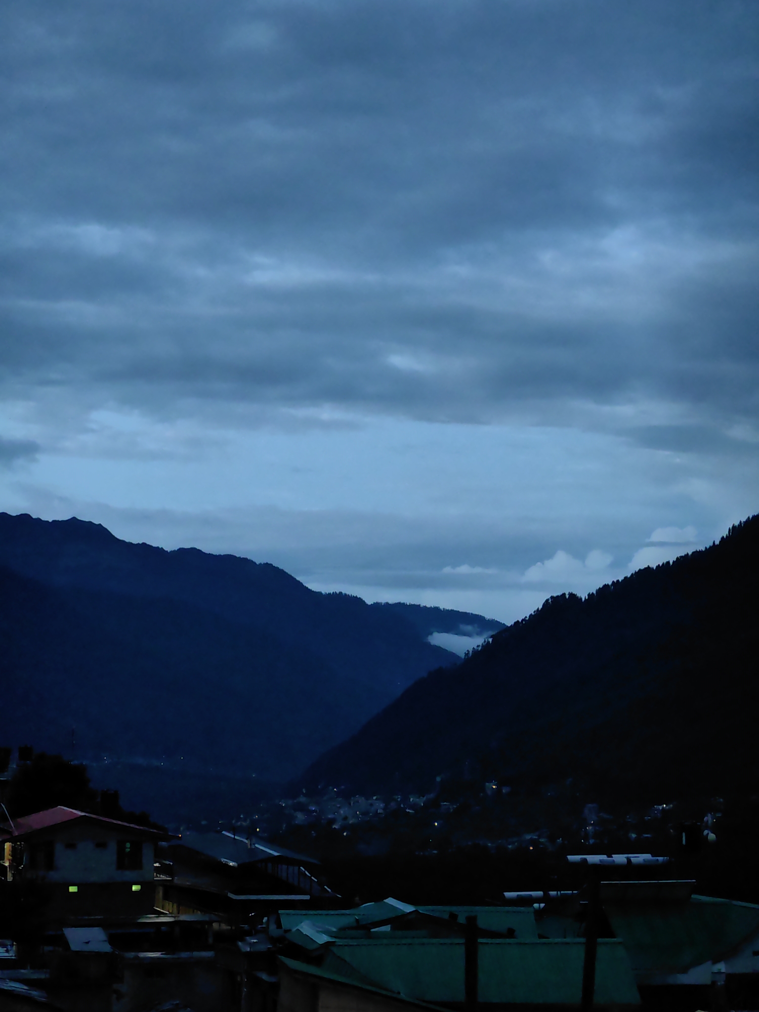 The image depicts a twilight scene in Vashisht, India, characterized by dark, moody skies filled with heavy clouds and the silhouettes of rolling mountains. In the foreground, several buildings with pitched roofs are visible, some appearing to be homes with windows illuminated by interior lights. The architecture suggests a village or small town setting nestled within the mountainous terrain. In the mid-ground, a distant settlement can be seen with scattered lights, suggesting a valley town or village. The overall atmosphere is serene and somewhat somber due to the low light conditions and overcast sky.