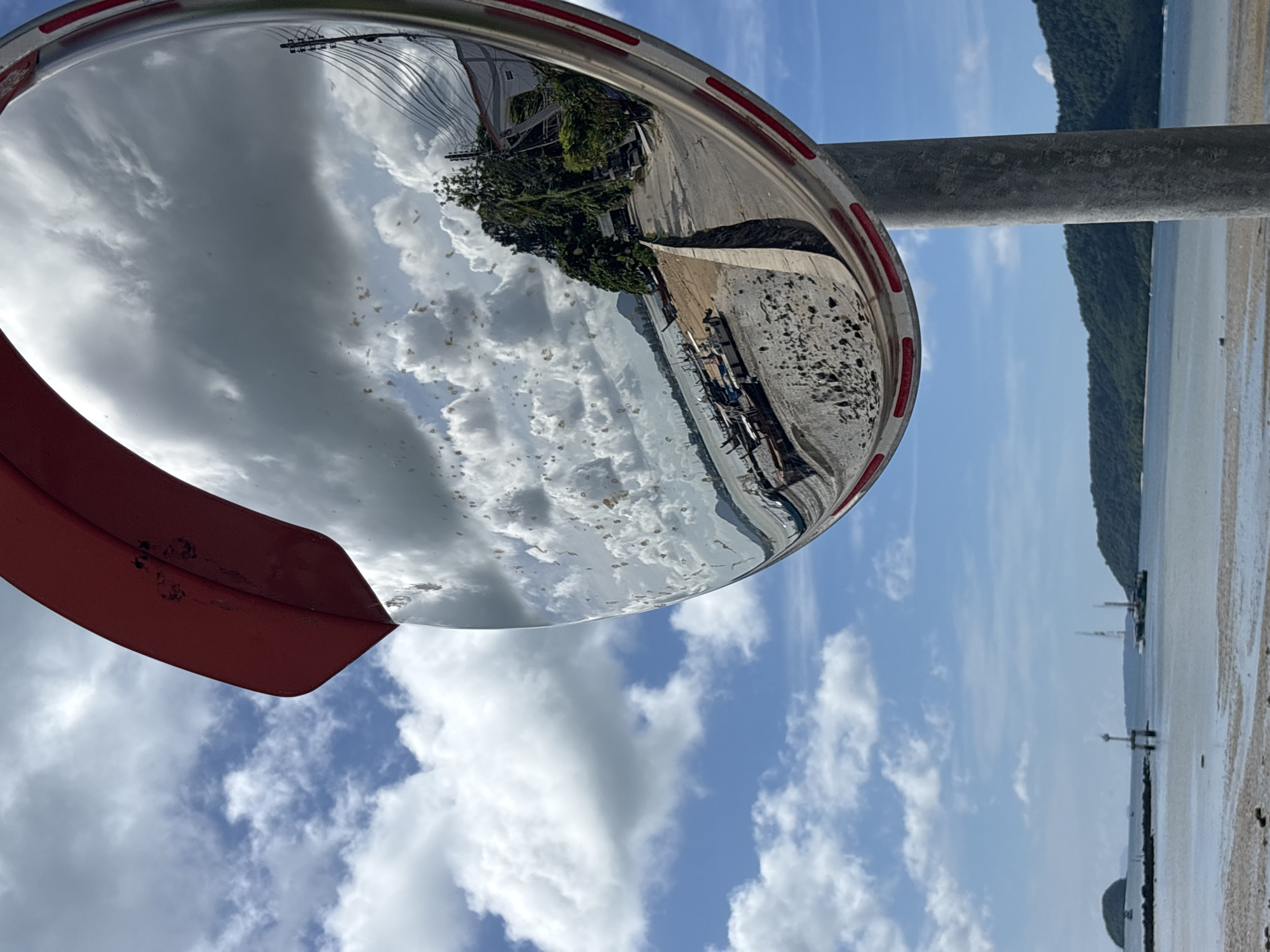 A convex traffic mirror, likely located near Rawai, Thailand, reflects a coastal scene. The mirror, framed in red and white, shows a cloudy sky and a view of boats pulled onto a sandy shore. A road curves away from the shore, bordered by a concrete wall, with buildings and trees visible on the right side of the reflection. In the background, beyond the mirror's reflection, a body of water stretches towards a tree-covered island and hills under a blue sky with scattered clouds. A lighthouse-like structure and some boats are visible in the water.