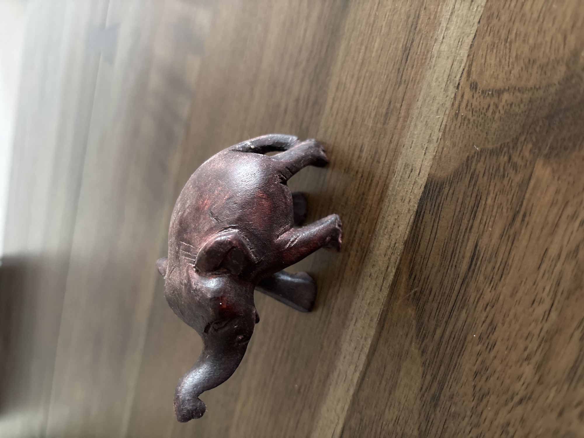 A carved wooden elephant figurine rests on a polished wooden surface. The elephant is dark brown, with visible grain and some red undertones, and is positioned facing left with its trunk raised. Its ears are small and tucked back, and its eyes appear to be small red beads. The wooden surface is a rich brown with distinct grain patterns, reflecting light and creating soft shadows. The background is out of focus, showing lighter tones that suggest an indoor environment. There is no indication of the city of Dubai or the United Arab Emirates.