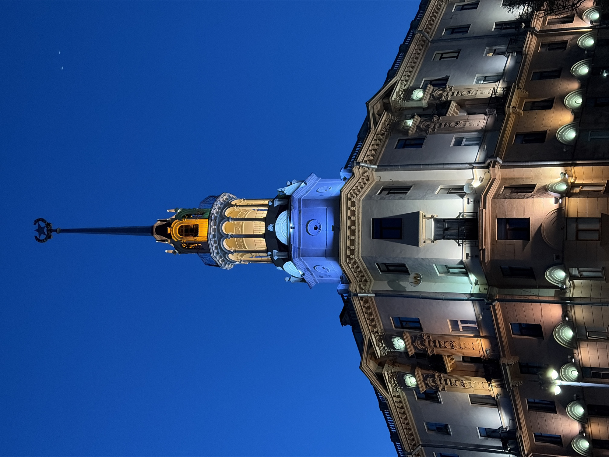 The image shows the iconic Stalinist Empire-style skyscraper at Nezavisimosti Square in Minsk, Belarus, illuminated against a dark blue twilight sky. The tower features a sharp spire topped with a star. The building's facade is adorned with decorative elements and illuminated by a mix of warm yellow and cool blue lights. The lighting highlights architectural details such as balconies, window frames, and sculpted ornamentation. There are no people visible in the image. The overall scene conveys a sense of grandeur and historical significance, typical of Soviet-era architecture.