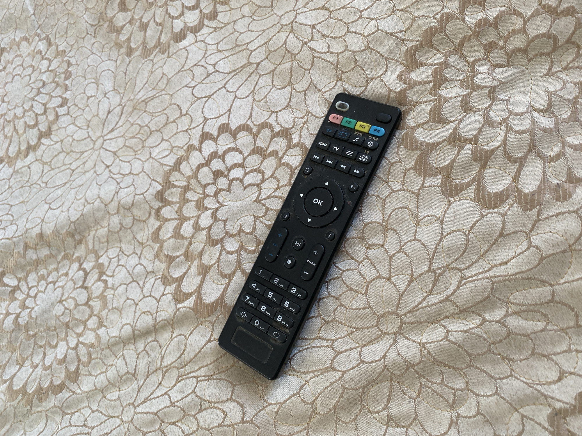 A black remote control with numerous buttons is positioned diagonally on a beige fabric with a floral pattern. The remote has numerical keys, navigation buttons, and function keys labeled F1, F2, F3, and F4. The fabric is textured with repeating large floral motifs in a lighter beige and brown hue. The image is taken from a top-down perspective. There are no people or animals visible. The scene is indoors, on a flat surface. The location of Chornomorsk, Ukraine is not discernible from the image.