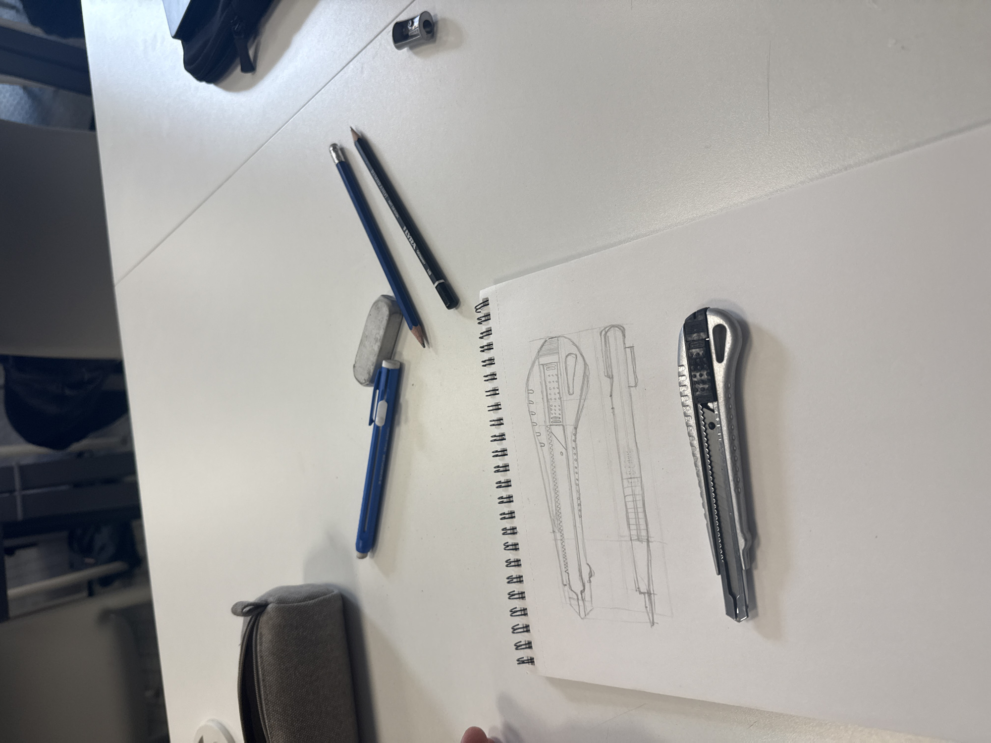 The image captures a white desktop workspace featuring various drawing tools and a sketchbook. On the left side of the table, two pencils are visible: one dark pencil with the text "REMBRANDT ART DESIGN LYRA Germany 10B" imprinted, and one blue pencil. Alongside them are a grey rectangular eraser, a small silver metal pencil sharpener, and a blue retractable utility knife. A grey zippered fabric pencil case is positioned towards the bottom left.
An open, spiral-bound sketchbook occupies the right side of the frame. It displays a detailed pencil drawing of a utility knife, rendered from multiple perspectives or views. Resting next to the sketchbook, a real silver-colored retractable utility knife with a serrated grip serves as the apparent subject of the drawing. The scene suggests an art or design activity, specifically a still-life drawing exercise. The lighting indicates an indoor, daytime setting. The provided location context is Milan, Italy.