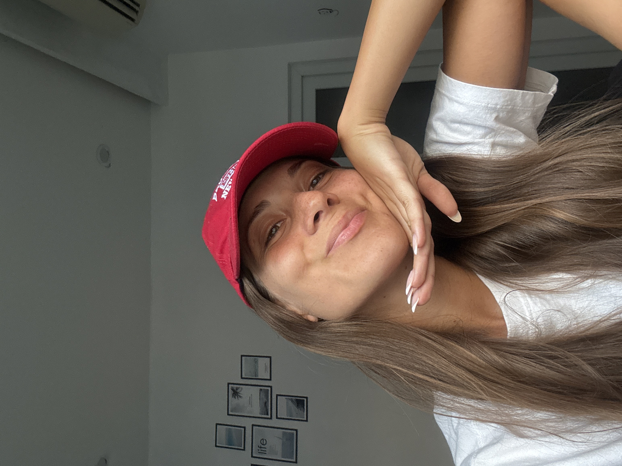 A woman wearing a red cap and a white t-shirt poses for a selfie. She rests her hand on her cheek, and her long brown hair falls to the side. In the background, there's a white wall with a collection of framed pictures depicting beach scenes and the word "life". A window is visible, allowing natural light into the room. An air conditioning unit is mounted high on the wall. No specific location in Đà Nẵng is ascertainable from the image.