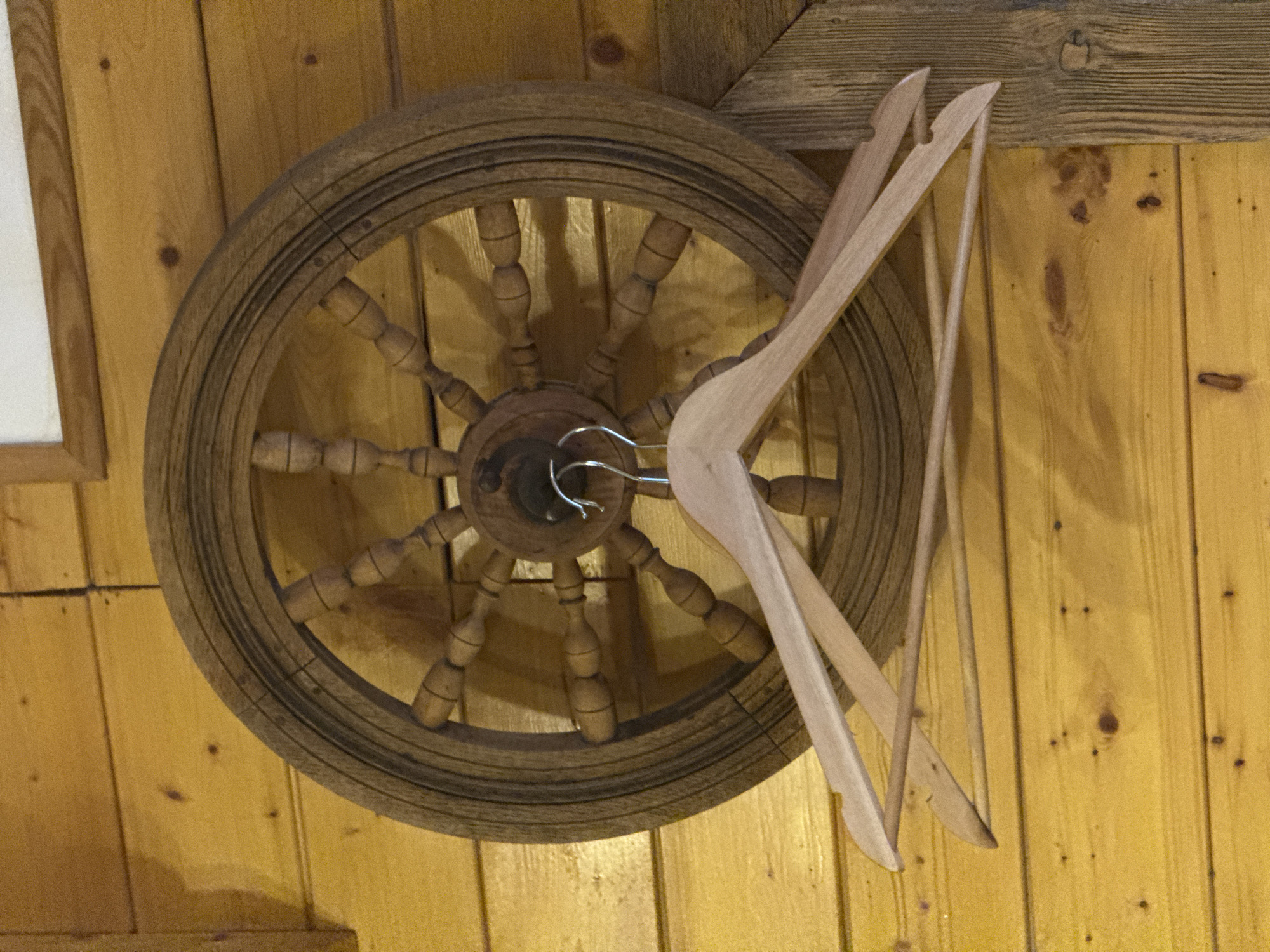 An antique wooden steering wheel, with a metal hook, hangs on a textured yellow wooden wall alongside two wooden coat hangers. The location is identified as the city of Ketoshi, Russia.