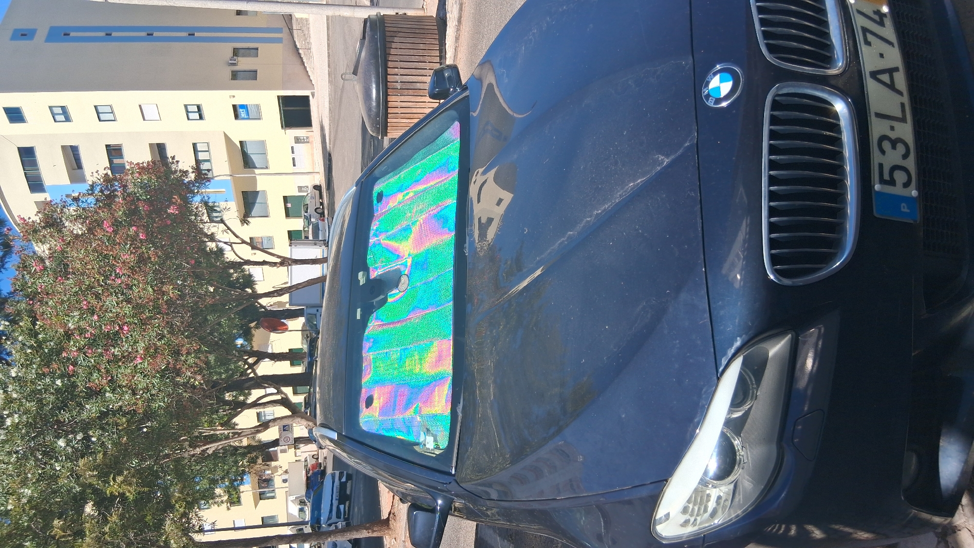 An exterior, eye-level, medium shot captures a dark-colored BMW sedan parked on a street in Setúbal, Portugal. The car faces the viewer, with its hood and front grille prominently displayed. A holographic sunshade is installed on the windshield, reflecting a rainbow of colors, creating a visually striking effect. The car's headlights are visible on the left, and the license plate, with the registration "53-LA-74", is clearly readable on the front bumper.
Behind the car, a modern, multi-story building with a light yellow facade and blue accents lines the street. Trees with green foliage and some pink blossoms are scattered in the background, partially obscuring the building. A dark, cylindrical trash receptacle with a wooden slatted exterior is positioned to the right of the car. The ground appears to be paved with a combination of asphalt and cobblestones. The sky is clear and blue, indicating a sunny day. No people are visible in the frame. The overall scene depicts a typical urban parking situation on a bright day.