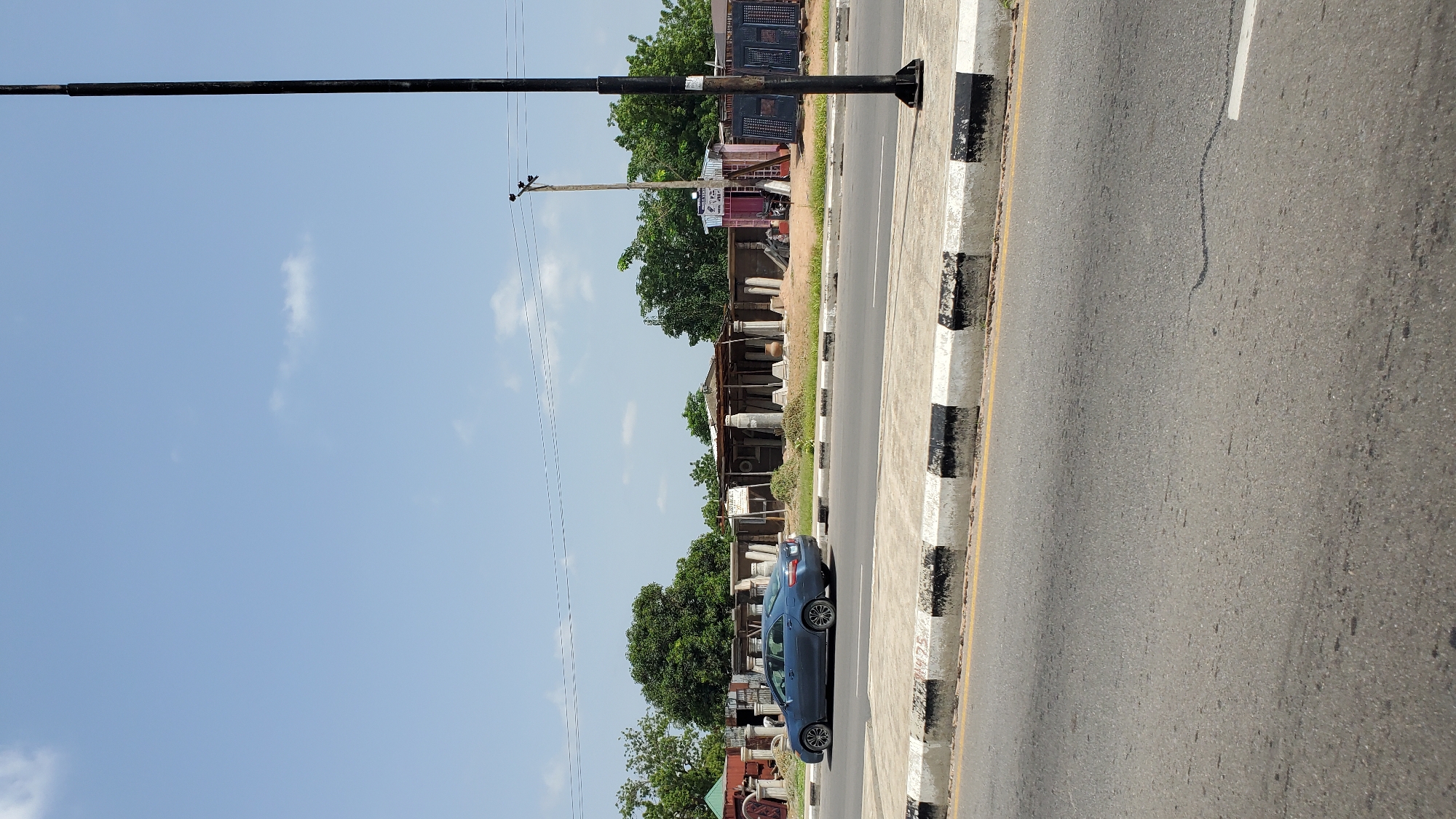 This is a low-angle, wide-shot photograph taken outdoors in daylight, likely in the late morning or early afternoon given the bright sky and the shadows. The image is oriented vertically, with the top of the frame showing a vast expanse of clear blue sky dotted with a few wispy white clouds. A thick, black, horizontal pole stretches across the upper portion of the image, obscuring some of the sky.
Below the sky, the scene transitions to a roadside setting. A black and white striped concrete curb runs along the edge of an asphalt road. The road itself is gray and shows some signs of wear and tear, with a faint white line marking the lane. To the left of the curb, there is a narrow strip of grass and then a collection of structures that appear to be shops or market stalls. These structures are characterized by corrugated metal roofs and open fronts, displaying various items for sale. Among the items visible are large, cylindrical stone or concrete columns, and what appear to be decorative architectural elements. Some of these are stacked and displayed outdoors. Further back, behind the stalls, are lush green trees.
A blue sedan is parked on the side of the road, partially visible and facing away from the camera. The car is the only discernible moving object in the frame. The overall atmosphere is one of a typical roadside market or commercial area in a warm climate. There are power lines and a utility pole visible, indicating the presence of infrastructure. One of the stalls has a sign with text that is too blurry to read. The scene suggests a busy commercial area, though there are no people clearly visible. The location context of Jalingo, Nigeria, points to a setting in West Africa.