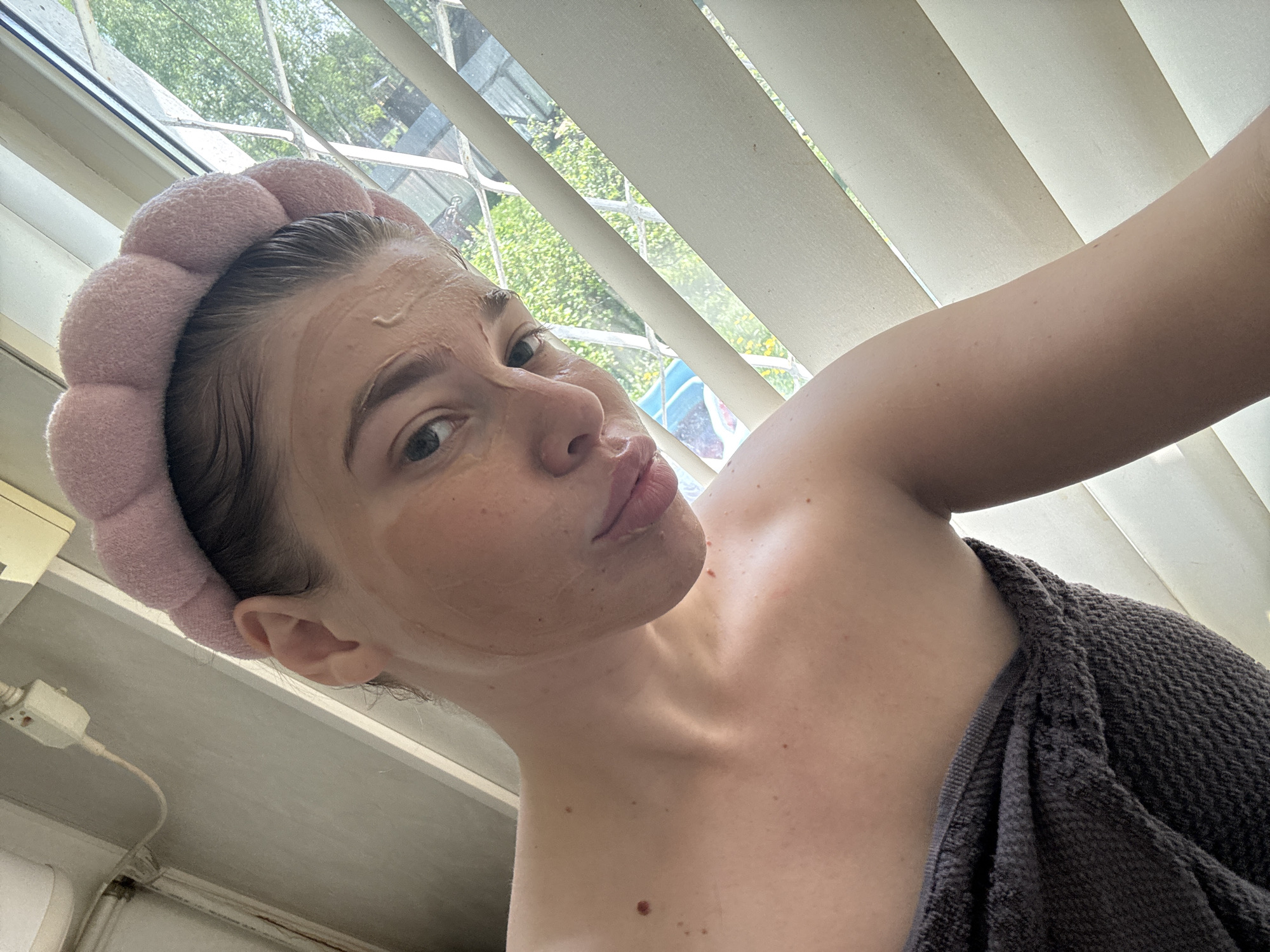 A low-angle, indoor selfie shows a fair-skinned young woman with a facial mask applied. She wears a pink, puffy headband and a dark gray towel draped over her shoulder. The background includes a window with closed vertical blinds and a view of trees and buildings. The location cannot be determined to be Мещёры, Russia based on this image.