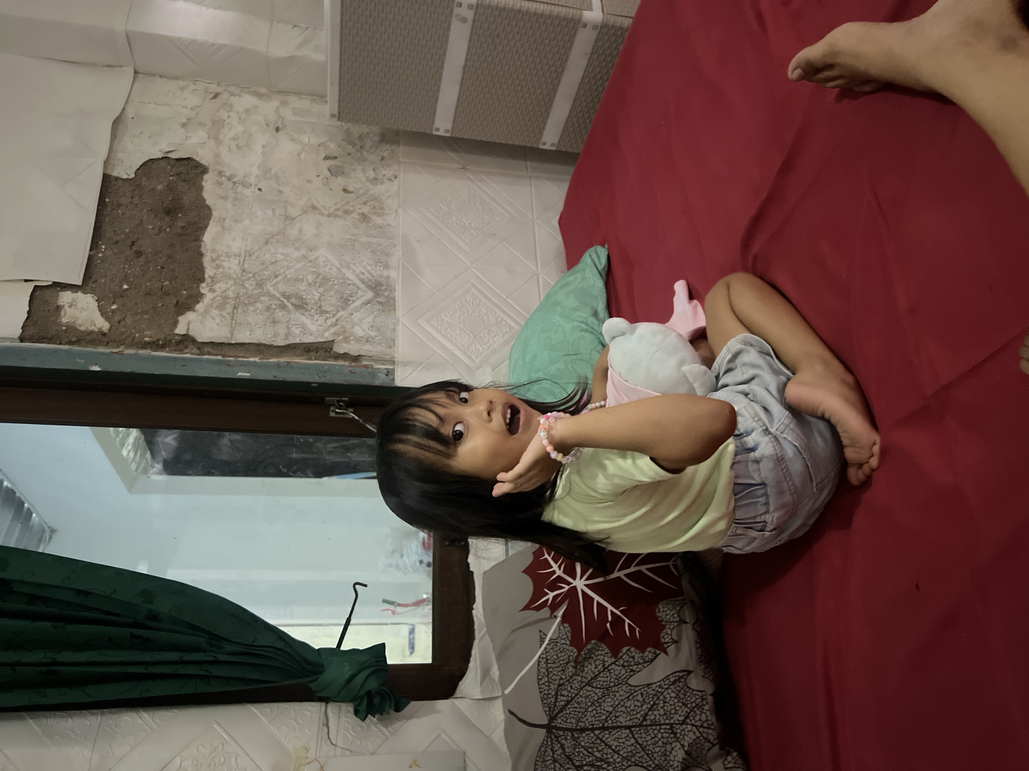 In this image, a young girl is lying on a red bed. She is wearing a light green t-shirt and denim shorts. The girl is holding a light blue stuffed animal and a pink toy. She has dark hair and is looking at the camera with a surprised expression. Her right hand is raised to her face, and she is wearing a beaded bracelet on her left wrist. Behind her, a teal pillow is visible, and a red blanket covers the bed.
The background of the image shows a wall with peeling paint and some textured tiling. To the left, a window with a dark frame reveals a glimpse of what appears to be an interior space with a green curtain. The scene is indoors, and the lighting suggests it could be daytime. The overall mood is playful and candid. The presence of the stuffed toys indicates the girl is likely engaged in a moment of leisure or play.