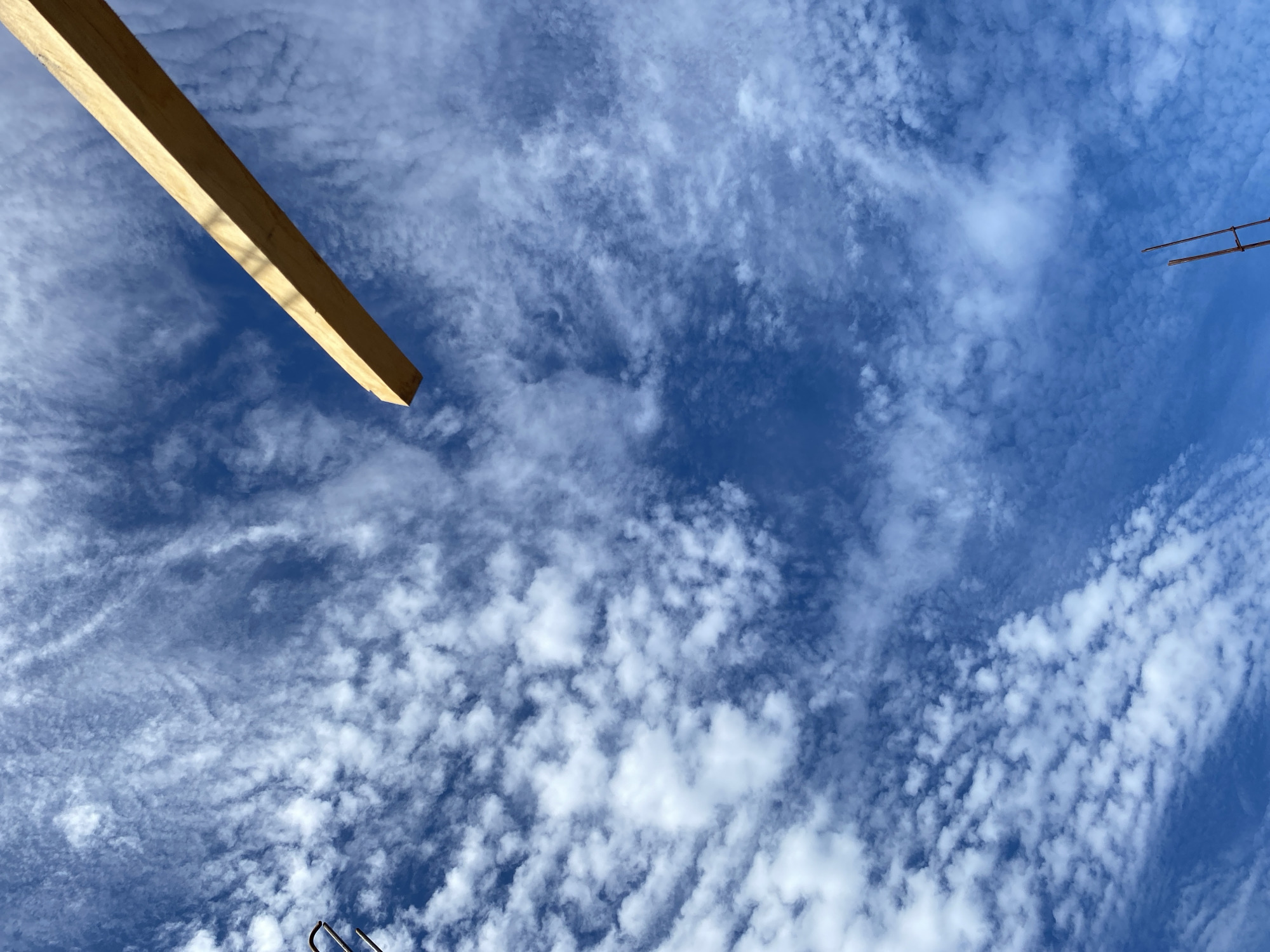 This is a low-angle, outdoor, full shot image looking up at a blue sky filled with scattered white clouds. A light brown wooden plank extends diagonally from the upper right corner towards the center of the frame. In the lower left portion of the image, the top of a metallic object, possibly rebar, is visible. At the bottom of the frame, two thin, dark poles stand vertically, one slightly to the right of the other. The scene appears to be in an outdoor construction area, possibly in Banda Aceh, Indonesia, given the context. There are no people visible in the image.