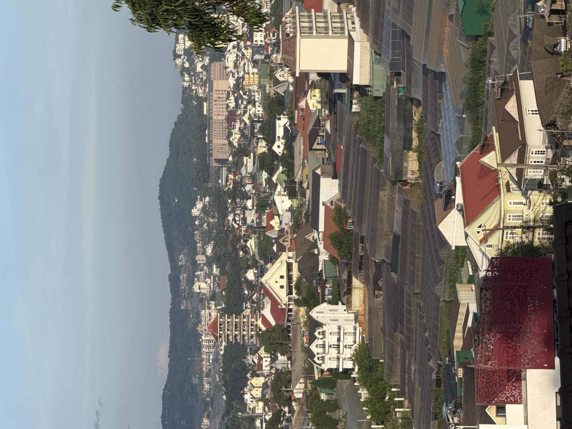The image depicts a sprawling cityscape from a high vantage point, likely in Lâm Viên - Đà Lạt, Vietnam, given the architectural style and landscape. The scene is dominated by numerous buildings of varying sizes and designs, nestled amidst lush green hills. In the foreground, a collection of greenhouses with translucent roofing stretches across the landscape, suggesting agricultural activity. Among the buildings, a prominent multi-tiered pagoda-like structure stands out, characterized by its red roof and intricate design. Several residential houses with pitched roofs, some painted in white or yellow, are interspersed throughout the scene. A tall, rectangular building with a light-colored facade and arched windows is visible on the right side of the frame. In the far distance, rolling hills covered in dense vegetation form the backdrop, meeting a hazy sky. The overall impression is one of a densely populated urban area integrated with the natural environment. No people are visible in the image.