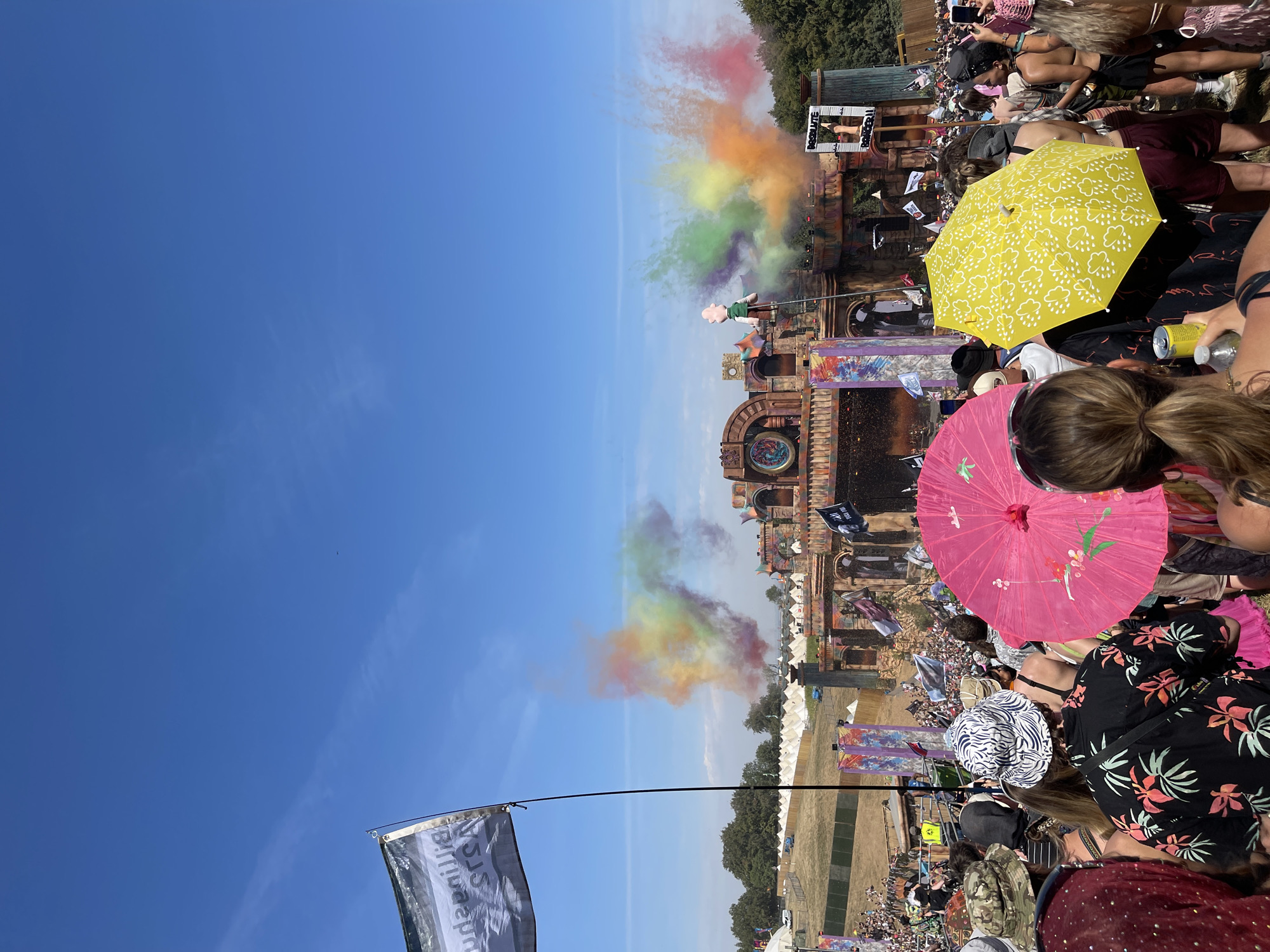 The image depicts a vibrant outdoor music festival with a large crowd gathered in front of an elaborate stage. The stage is ornately decorated with what appear to be ancient architectural elements and is adorned with banners and flags. Colorful smoke plumes erupt from the stage area, creating a visually dynamic spectacle against a clear blue sky.
In the foreground, the backs of several festival-goers are visible. They are dressed in casual, summery attire, with some individuals holding colorful umbrellas, one pink and one yellow. People are holding drinks and phones, suggesting active engagement with the event. To the left, a black and white flag with text is partially visible, extending into the upper left corner of the frame.
The background reveals a large field with numerous white tents, indicating a camping area associated with the festival. Beyond the tents, trees and a gently sloping hillside can be seen. The overall scene suggests a lively and energetic atmosphere at a daytime festival. The location is identified as the city of Winchester, United Kingdom.
