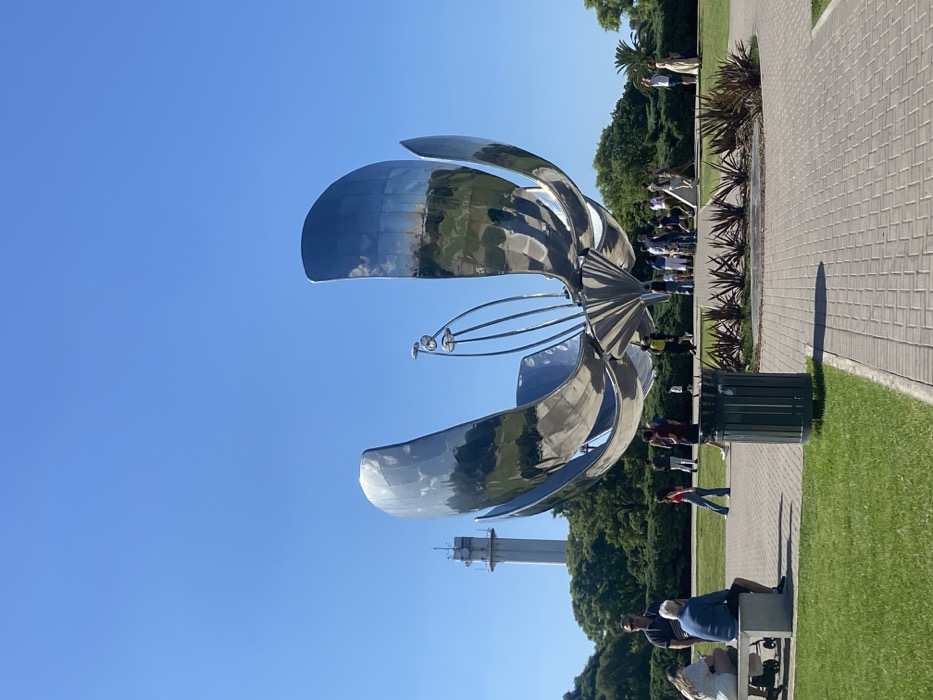 A large, modern, metallic flower sculpture dominates the image, with its reflective petals mirroring the surrounding park. People mill around the sculpture and line a paved path. A tall tower-like structure is visible in the background. The sky is clear and blue. Green grass and lush trees create a vibrant park setting.