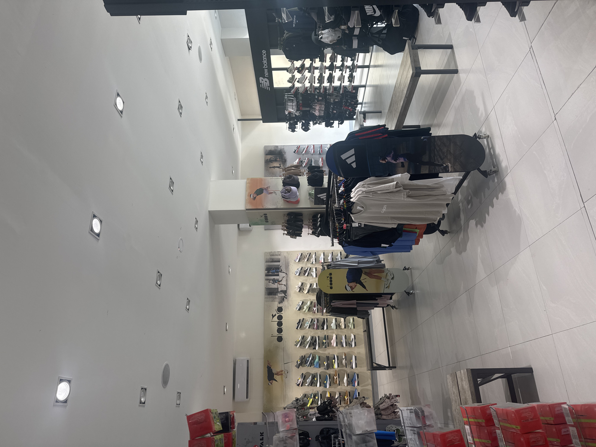 The image captures the interior of a retail store in North Kuta, Indonesia, likely specializing in athletic apparel and footwear. The space is brightly lit with numerous recessed lights in the white ceiling. The walls are also white, creating a clean and modern aesthetic.
Merchandise is displayed on racks and shelves along the walls. Brand signage for "New Balance" is visible. There are displays of shoes, clothing, and accessories. The aisles are clear, suggesting a well-organized and maintained retail environment.
No people are visible in the frame.