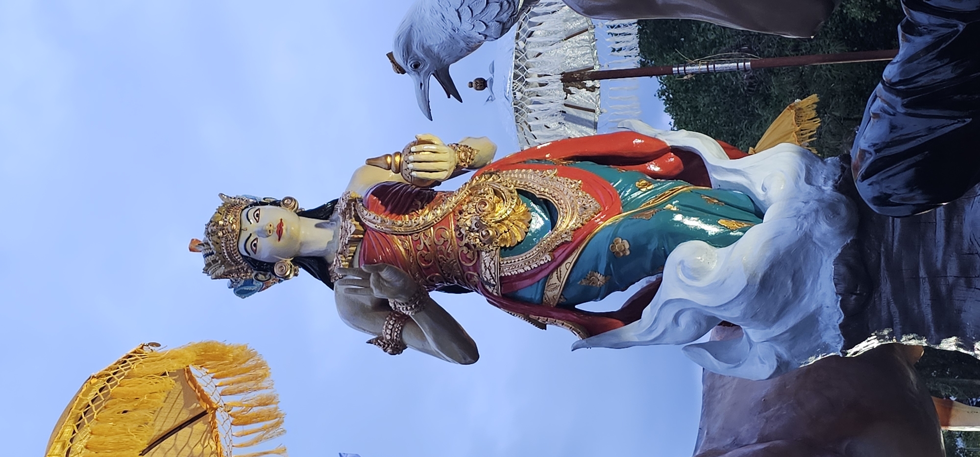 This image captures a large, intricately sculpted statue, likely depicting a Balinese Hindu deity or mythological figure, set against a clear, light blue sky. The central subject is a female figure presented in a graceful, reclining pose. She features pale skin, dark hair, and a serene expression with red lips and dark, almond-shaped eyes. Her elaborate attire includes a golden crown-like headpiece and multiple pieces of ornate jewelry on her arms and neck. Her garments are vibrantly colored in rich reds, greens, and gold, adorned with intricate patterns and golden embellishments. The statue's base is sculpted to resemble swirling white clouds.
Above and behind the figure's head, part of a large, white bird-like creature, possibly a mythical Garuda, is visible, extending its head downwards. Directly behind the figure's torso, a white ceremonial umbrella (*tedung* or *payung*) with white fringes can be partially seen, its shaft extending upwards. In the lower-left foreground, another yellow ceremonial umbrella is partially visible, indicating a decorative or ceremonial context. To the far right, a dark, partially obscured large structure or base provides a backdrop.
The scene takes place outdoors under a bright, clear sky, suggesting daytime or late afternoon. The detailed craftsmanship, traditional attire, and ceremonial umbrellas are characteristic of Balinese artistry and culture, consistent with the provided location context of Denpasar, Indonesia. This statue is likely part of a religious or cultural display, such as an *ogoh-ogoh* for Nyepi (Day of Silence) celebrations or a permanent temple ornament. No explicit activity is shown, as the statue is static. There is no visible text in the image.