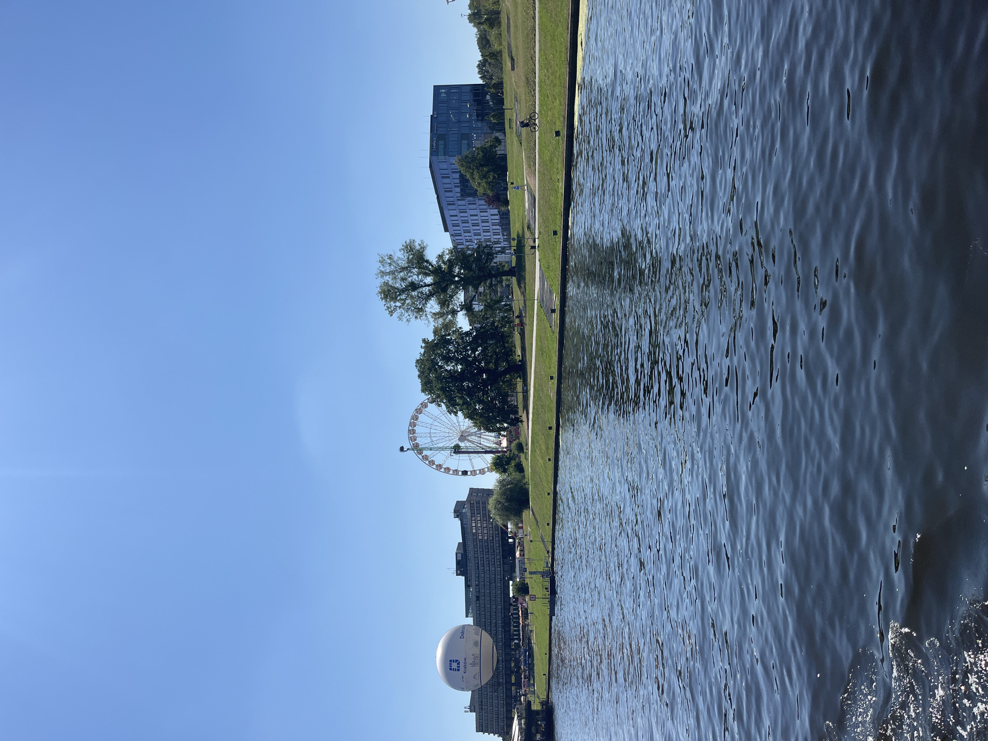 This is a landscape photograph taken from a body of water, likely a river, looking towards a city skyline on a clear day. The foreground is dominated by the dark blue water, with ripples and some white foam visible on the left.
In the midground, a grassy embankment runs along the water's edge. On this embankment, there are several notable structures. To the left, a large white hot air balloon is prominently displayed, with text and a logo visible on its side, possibly advertising a company or event in Krakow. Behind the balloon, a long, multi-story building with many windows stretches across the scene. Further to the right, a Ferris wheel stands tall, its circular structure and spokes clearly defined against the sky. Several trees are scattered across the embankment, with a few larger ones towards the right side of the image.
In the background, more modern buildings are visible, including a tall, rectangular building with many windows and a distinct blue-tinted facade. A person is seen cycling on a path along the grassy area in front of this building. The sky above is a clear, bright blue, with no clouds present. The overall impression is of a vibrant urban scene situated along a waterfront, under fair weather conditions. The presence of the hot air balloon with "Krakow" visible on it strongly suggests the location is Krakow, Poland.