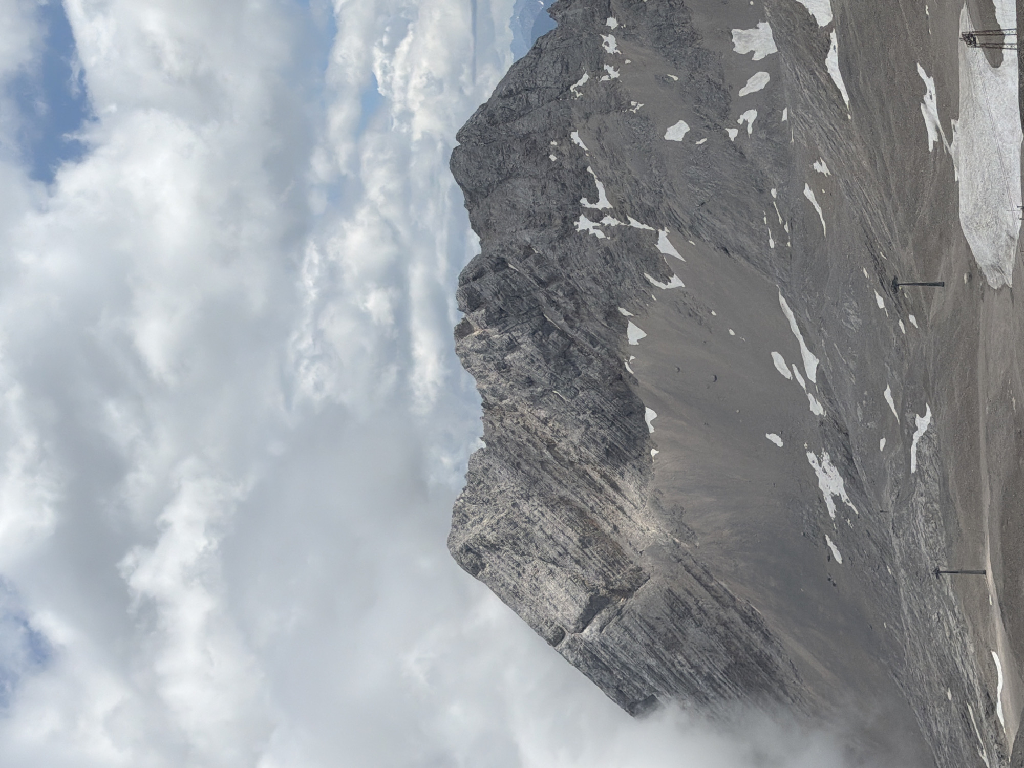 The image depicts a majestic mountain landscape under a cloudy sky. In the foreground, a barren, rocky slope is visible, interspersed with patches of snow. Two vertical structures, possibly ski lift towers, are present on the slope. The mountain itself is characterized by steep, rugged cliffs with visible geological strata. Clouds obscure parts of the mountain and extend into the sky. The scene is a natural, high-altitude environment, likely a mountain range. Considering the context of Garmisch-Partenkirchen, Germany, this view strongly resembles the Zugspitze massif, Germany's highest peak.