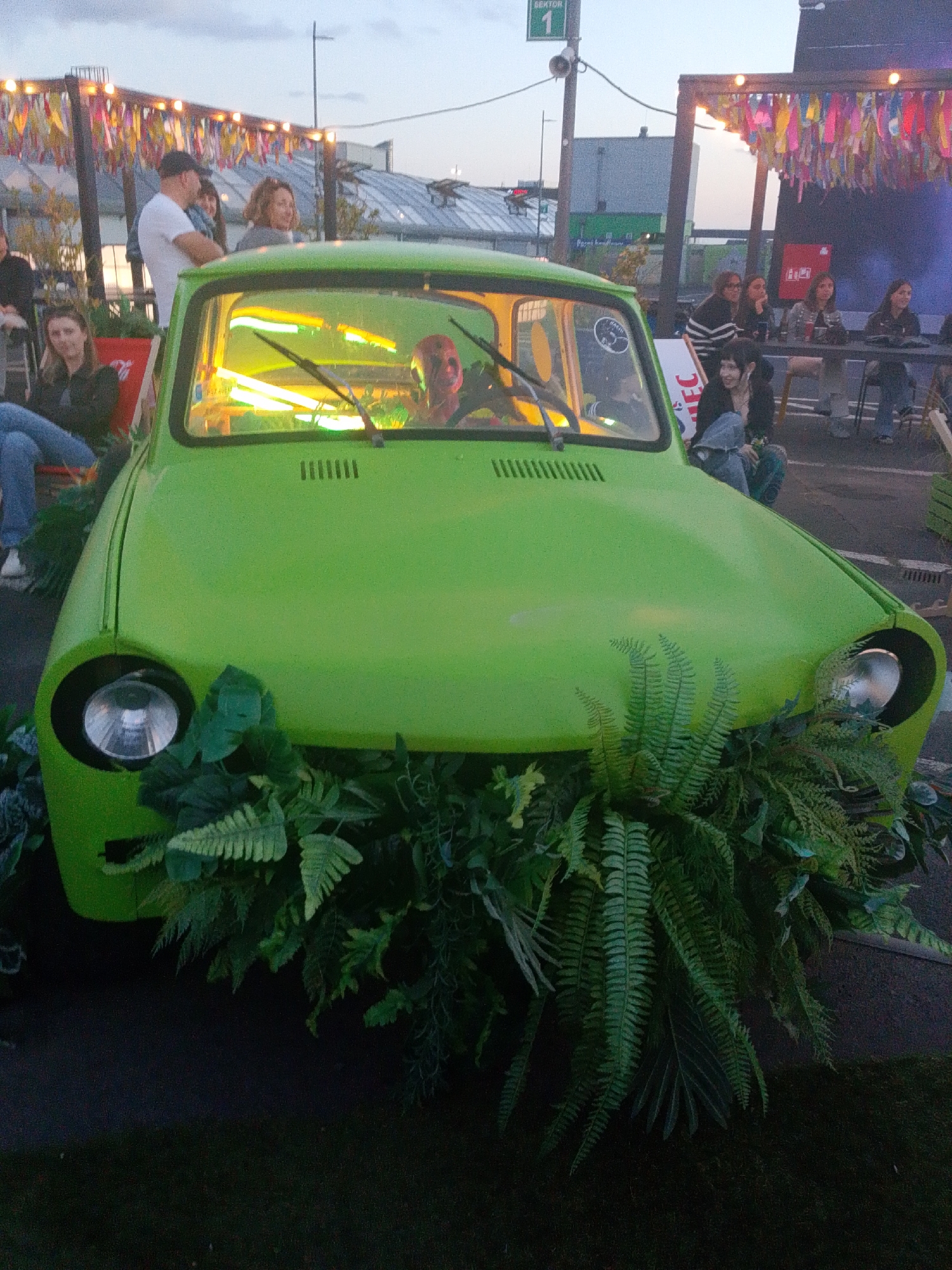A bright green, vintage car is parked outdoors, its front adorned with lush green ferns and foliage. Inside the car, illuminated by yellow neon lights, a person with their face painted red, possibly a clown or performer, is visible behind the steering wheel. In the background, a group of people are gathered around tables, seated on chairs beneath a canopy decorated with colorful streamers. One person stands near the car, wearing a white t-shirt and a black cap. The scene suggests an outdoor event or festival. The location is identified as the Wrocław Congress Center (Centrum Kongresowe Wrocław) in Wrocław, Poland, as indicated by a sign in the background reading "SEKTOR 1".