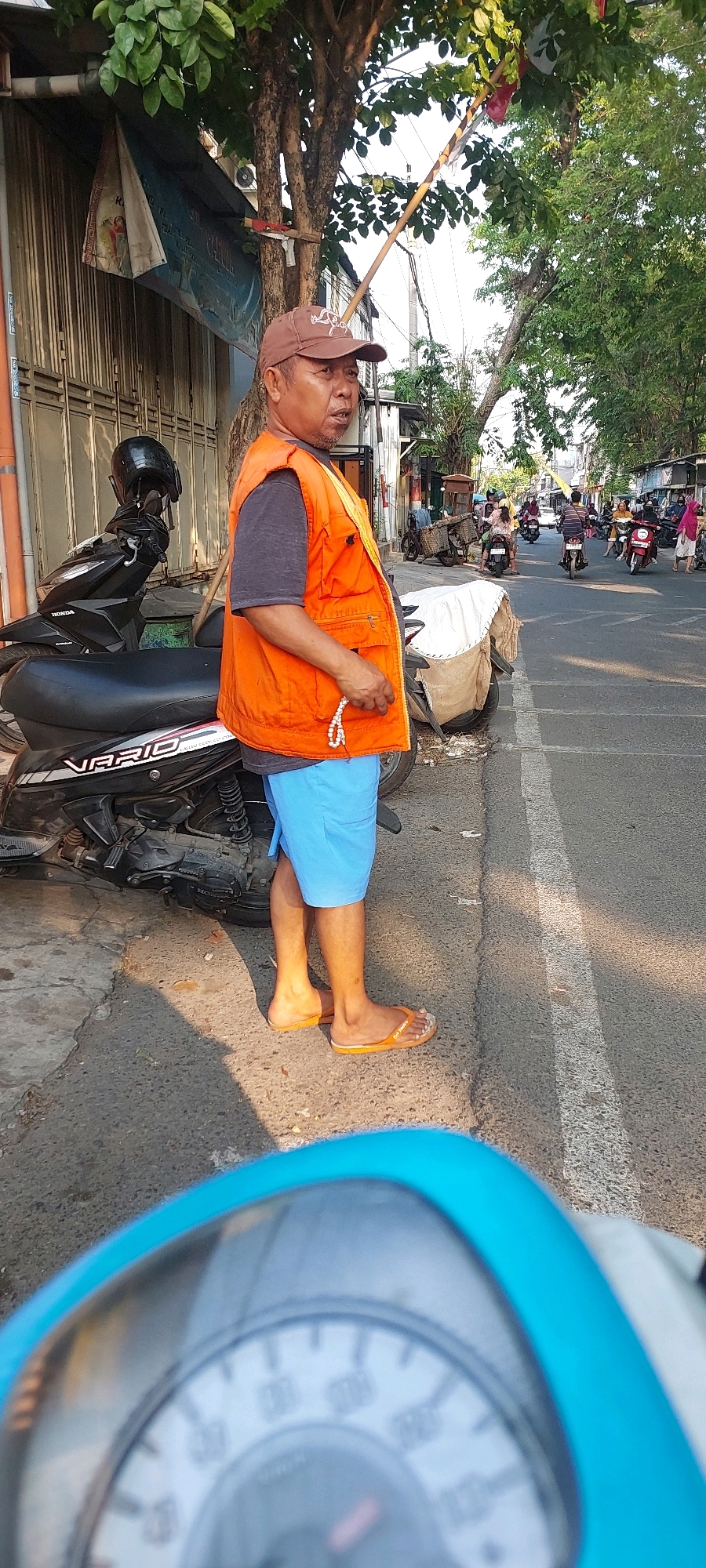 In Semarang, Indonesia, a man wearing an orange vest and blue shorts stands on a sidewalk next to a parked black scooter. He is looking towards the right of the frame, holding a string of white prayer beads in his right hand. Behind him, a street with traffic is visible, including several motorcycles and a woman walking. The man is standing in front of a building with a metal shutter and a tree with green leaves overhead. The ground is paved with asphalt and concrete. The overall scene depicts a typical street view in a bustling urban environment.