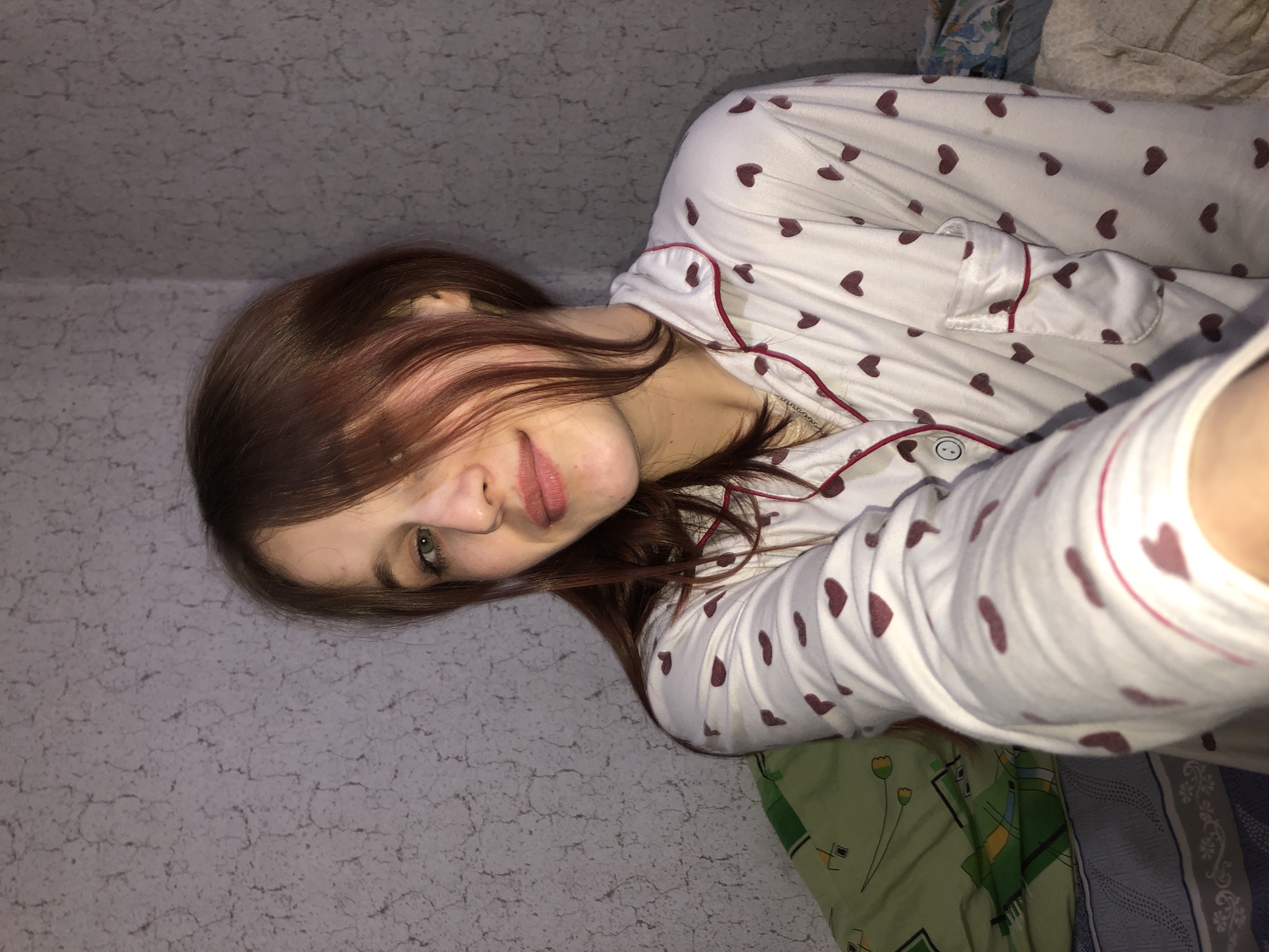 A young woman lies on a bed in an indoor setting. She is wearing a white pajama top with red heart patterns. Her hair is light brown, partially covering her face. The bed has green and blue patterned sheets. The walls appear to be a light grey color with a textured surface. The location is identified as Фоки, Russia.