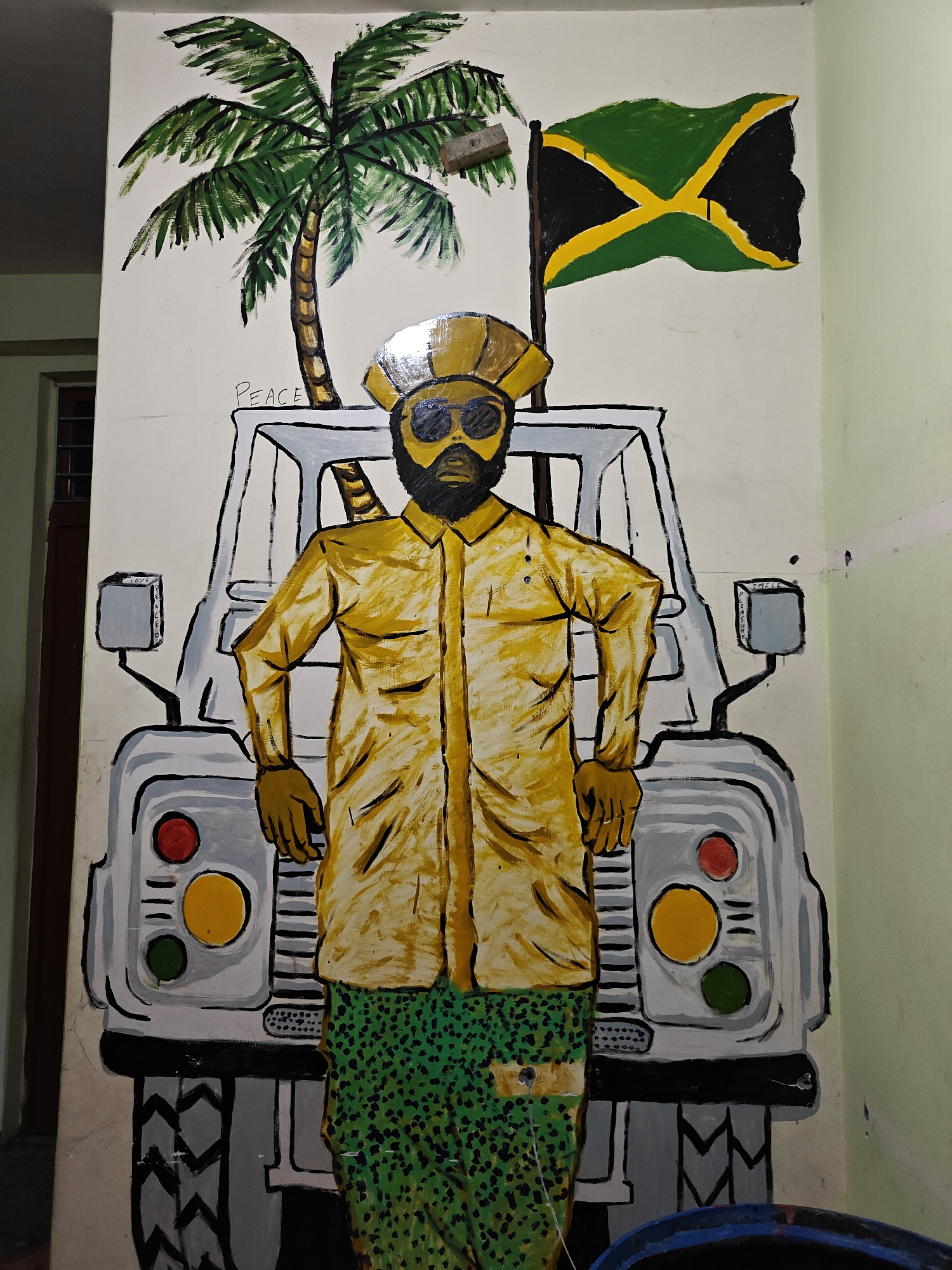 This image captures a vibrant mural painted on a white wall. The central figure is a stylized, dark-skinned man with a full black beard, wearing large round dark sunglasses and a golden-yellow head covering. He is dressed in a long-sleeved golden-yellow shirt and green pants with a black speckled pattern, standing in a confident pose with his hands on his hips.
Behind the man is a light grey, boxy vehicle, reminiscent of a classic Land Rover Defender. It features round headlights in red, yellow, and green on the front, and a red taillight on the rear. The vehicle's side mirrors are also depicted. To the left of the scene, a large green palm tree adds a tropical element. On the right, a Jamaican flag, with its distinctive black, green, and yellow diagonal cross design, waves from a pole.
The mural incorporates text with positive messages. "PEACE" is written in cursive above the vehicle near the palm tree. On the left side mirror of the vehicle, "LOVE" is inscribed vertically. The right side mirror displays "SMILE" horizontally and "PLEASURE" vertically below it. The painting, found in Manikaran, India, creates a static scene with strong cultural cues pointing to Jamaica or a reggae theme.