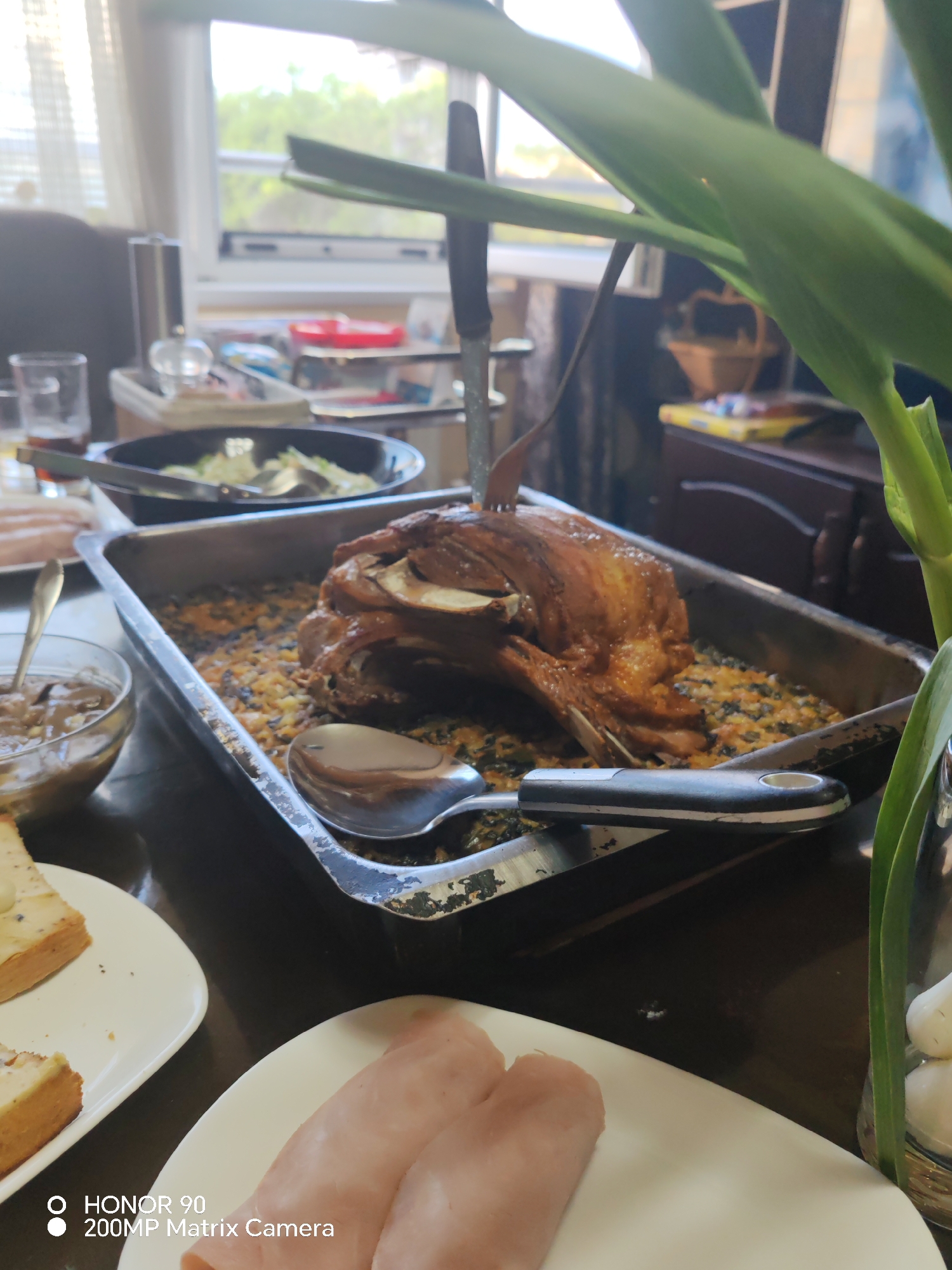 A table is set with a meal in what appears to be a home setting. A roasting pan contains a cooked meat dish with stuffing, a serving fork and a spoon are present. There's a bowl of mushroom sauce nearby, as well as plates of ham and toast. A salad bowl is positioned further back. Plant life in a glass jar is in the foreground. The scene takes place indoors, in daylight. Window is visible. It is not possible to identify if the scene takes place in Sofia, Bulgaria.