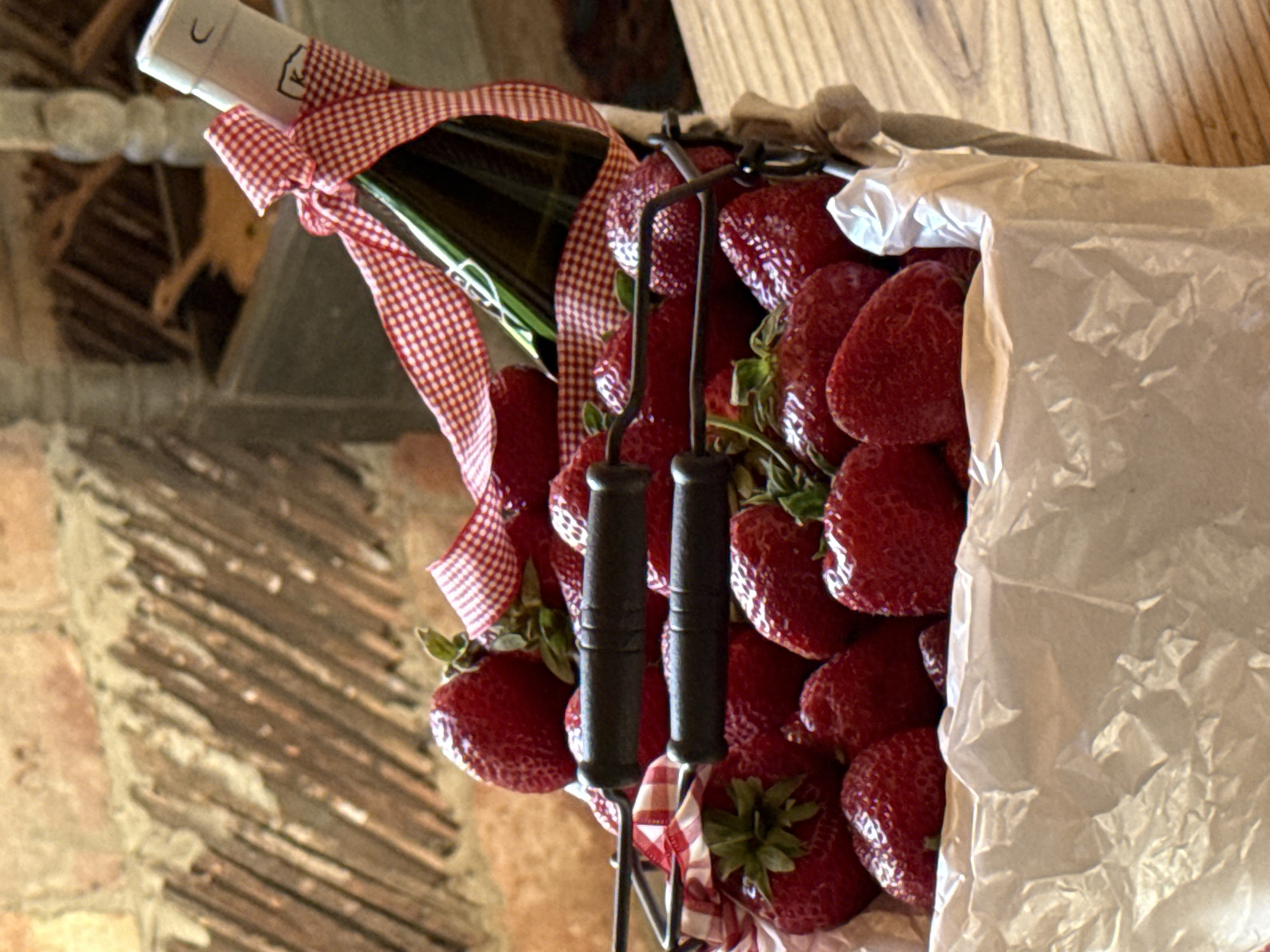 In Alagir, Russia, a dark metal basket filled with ripe strawberries and lined with white crumpled paper sits on a wooden surface. The basket has two dark handles. A green bottle, possibly containing champagne or wine, is placed among the strawberries and is adorned with a red and white checkered ribbon tied around its neck. The background features a rustic brick wall with wooden planks.