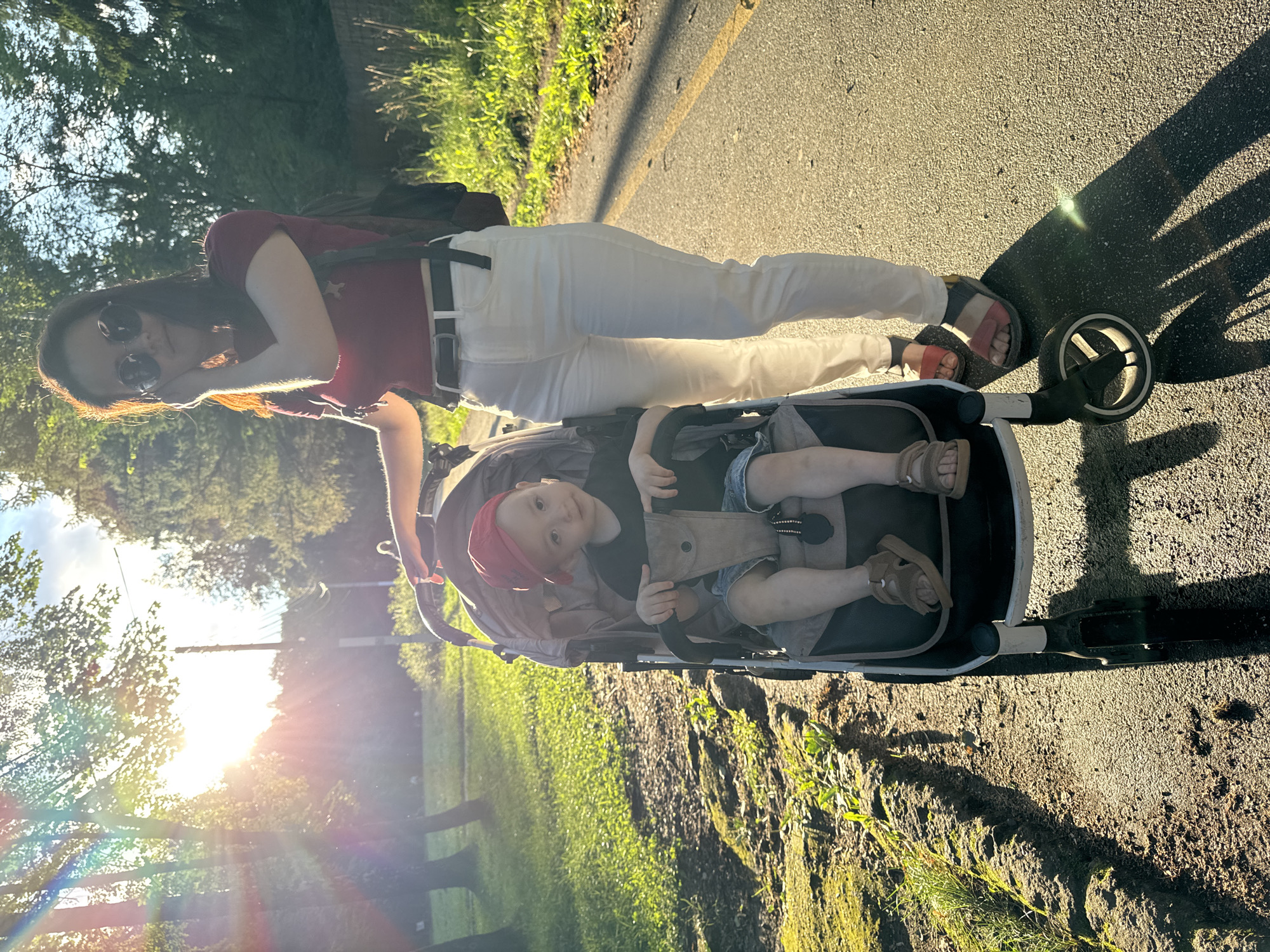 A woman stands next to a stroller containing a young child. The woman is wearing a maroon t-shirt, white pants, and sandals. She has sunglasses on and her hand is near her ear. The child is wearing a red hat and a black t-shirt. The stroller is grey and has a canopy. They are on a paved path in a park with trees and grass. The sun is shining brightly, creating lens flares and casting long shadows. The image was taken in Izhevsk, Russia.