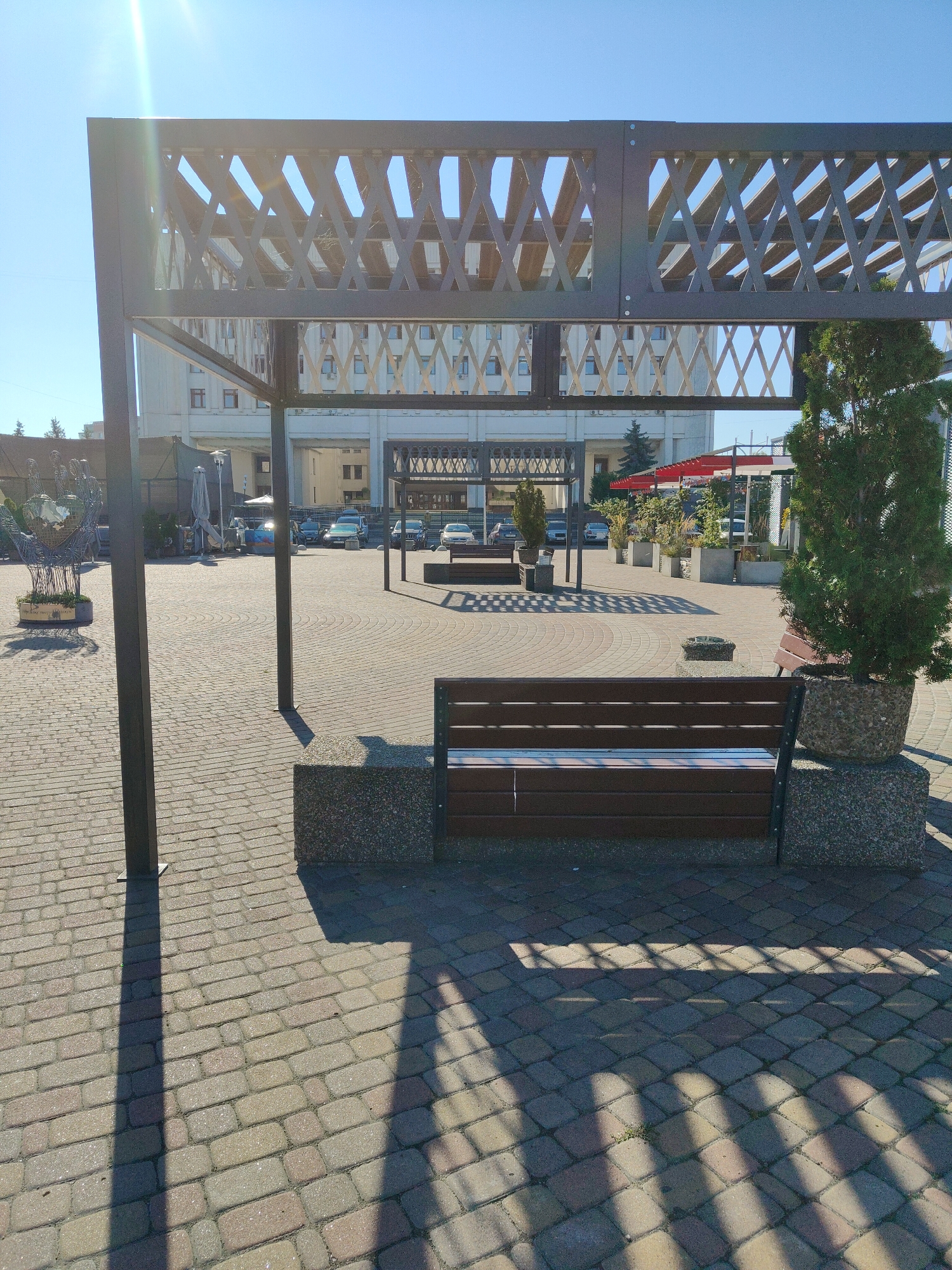 The image shows an outdoor seating area with a modern pergola structure in the foreground. The ground is paved with interlocking bricks, forming a patterned surface. Beneath the pergola, there is a dark brown wooden bench with a concrete base. To the right of the bench is a tall, potted evergreen tree. In the background, a light-colored building with multiple windows and balconies is visible. Cars are parked in front of the building, and there are several planters with greenery. A decorative metal sculpture resembling hands holding a globe is located to the left of the frame. The scene appears to be a public space, possibly a courtyard or plaza. Based on the architecture and overall aesthetic, this location could be in the city of Cherkasy, Ukraine, possibly near a business or hotel establishment. The lighting suggests it is a sunny day, casting distinct shadows from the pergola onto the paved ground. There are no people visible in the image.